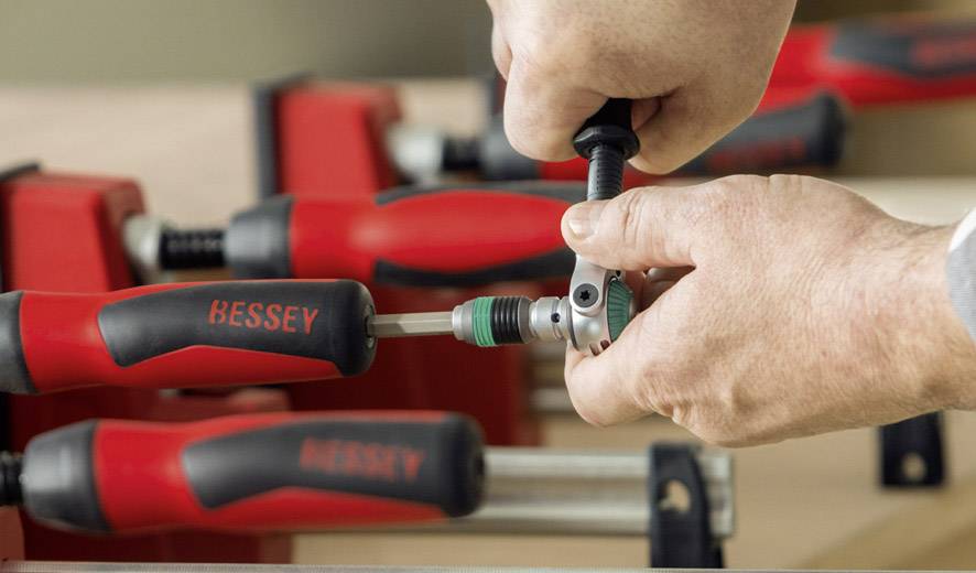Eine Person bedient eine rote Schraubzwinge mit der Aufschrift 'BESSEY'. Im Hintergrund sind weitere Schraubzwingen sichtbar.
