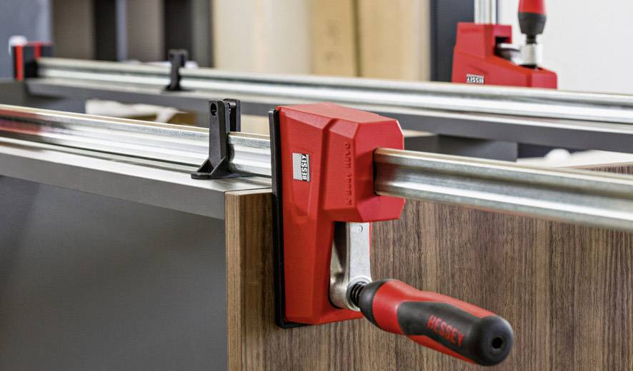 Eine rote Schraubzwinge hält zwei Holzplatten zusammen. Sie befindet sich in einer Werkstattumgebung und wird für Präzisionsarbeiten genutzt.