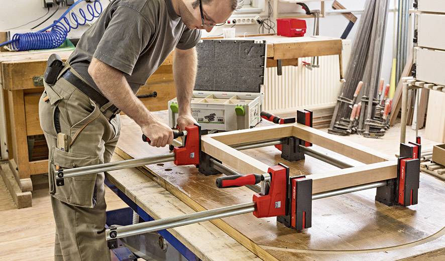 Ein Handwerker befestigt ein Holzgestell mit Schraubzwingen auf einer Werkbank in einer Werkstatt. Werkzeuge und Materialien sind im Hintergrund sichtbar.