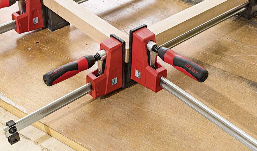 Zwei rote Zwingen fixieren Holzbalken auf einer Werkbank, ermöglichen präzises Arbeiten bei der Holzbearbeitung.