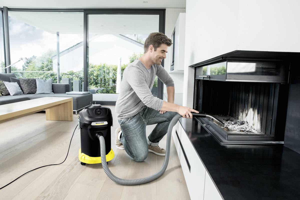 Eine Person kniet vor einem modernen Kamin und benutzt einen Aschestaubsauger, um Asche zu entfernen. Das Wohnzimmer ist geräumig und hell erleuchtet.