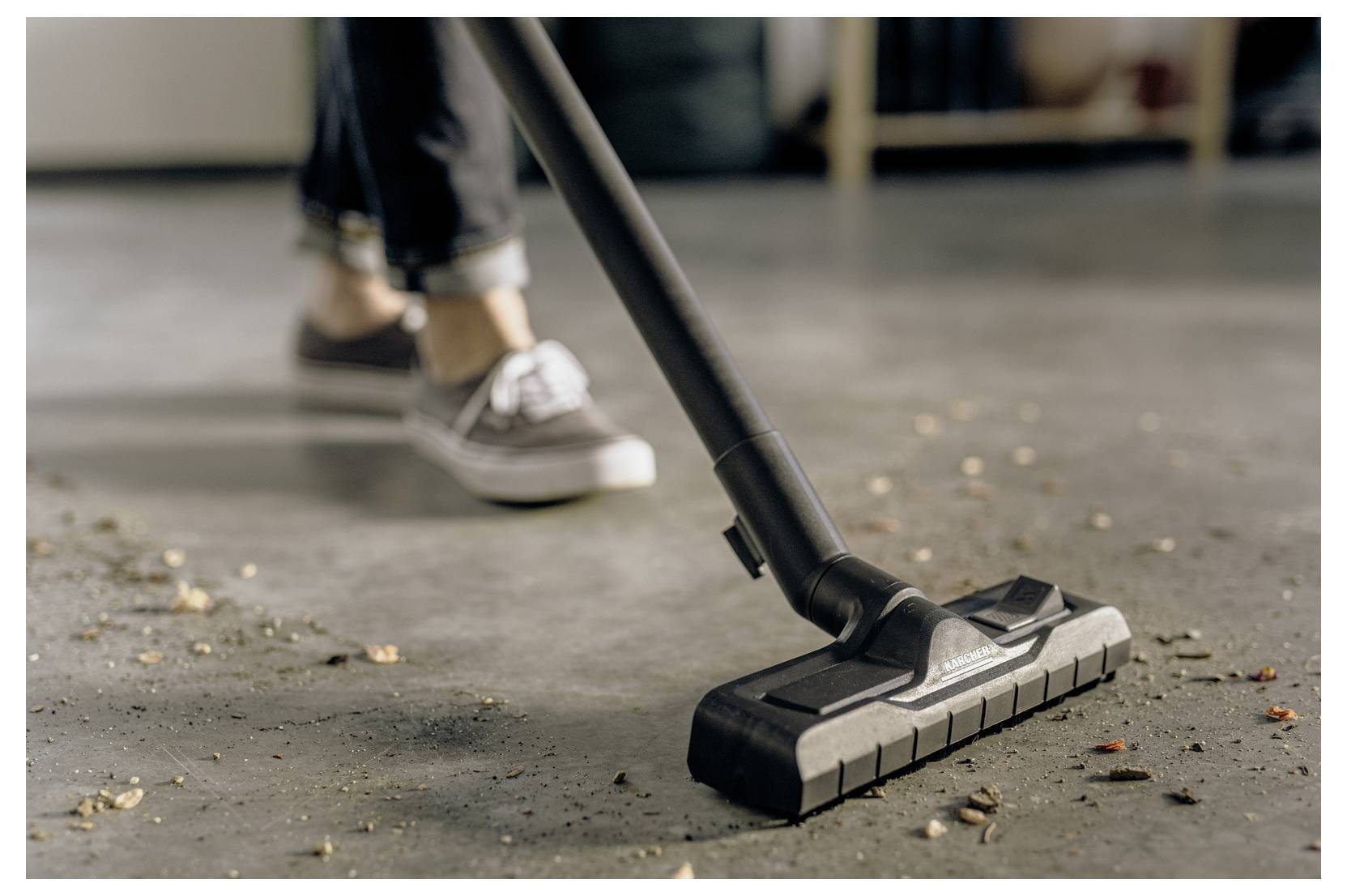 Eine Person, die einen Boden mit verstreutem Unrat saugt und sich dabei auf den Saugkopf konzentriert, trägt beiläufige Schuhe und Jeans.