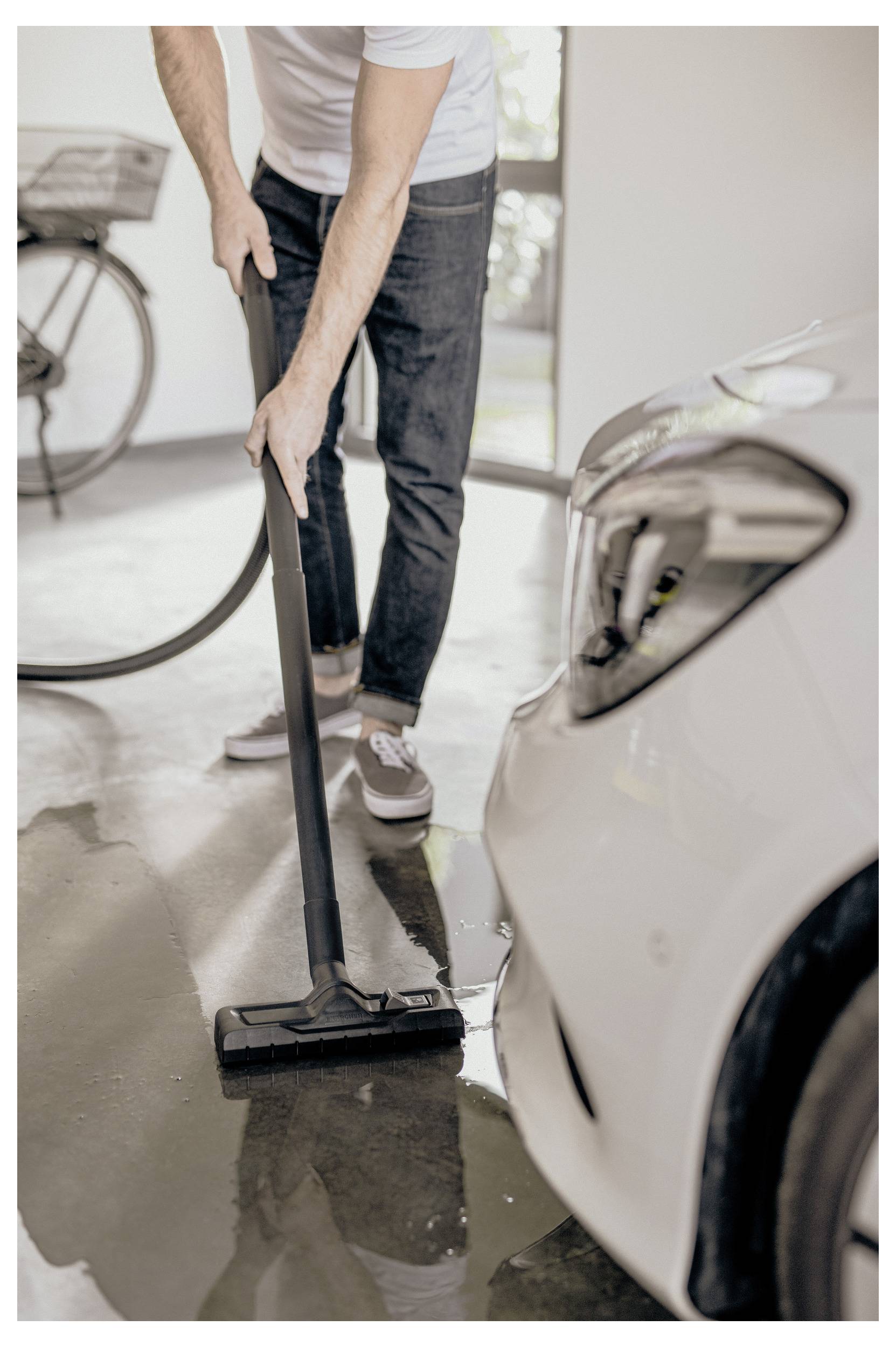 Eine Person verwendet einen Staubsauger, um einen nassen Garagenboden neben einem Auto zu reinigen und damit eine Reinigungs- oder Wartungsaktivität durchzuführen.