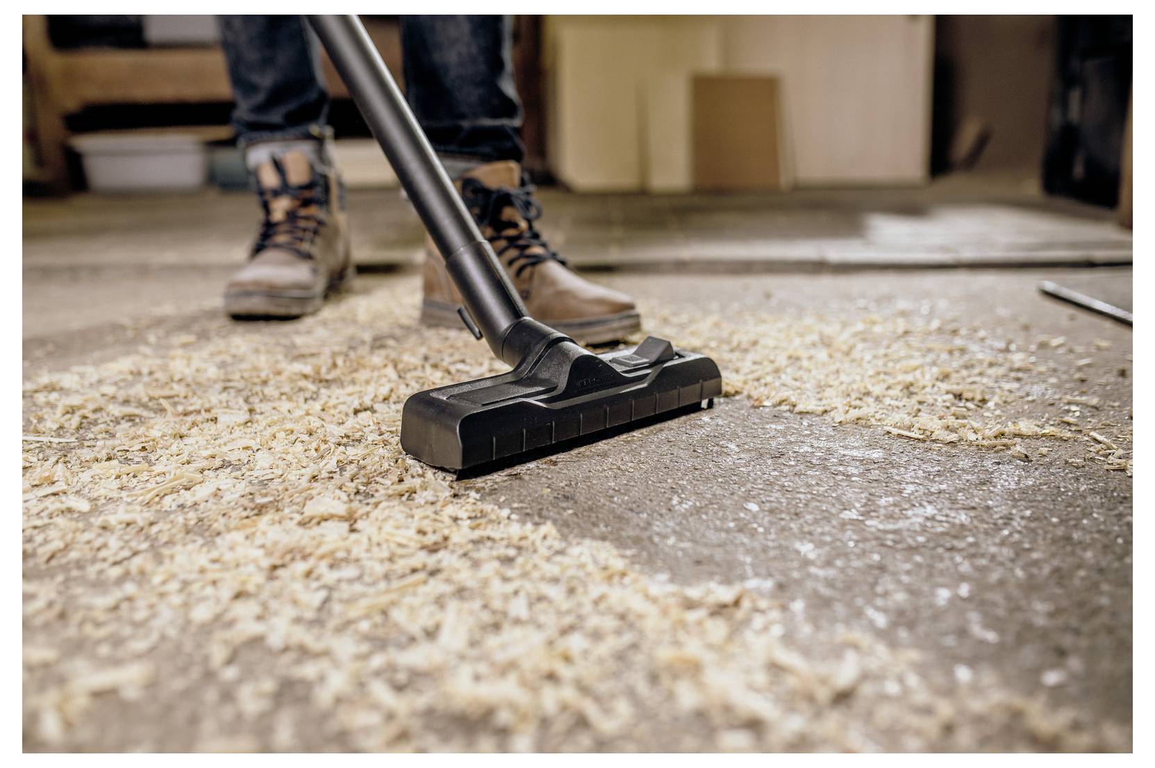 Eine Person benutzt einen Staubsauger, um Holzspäne von einem Werkstattboden zu entfernen, trägt Arbeitsschuhe und Jeans.