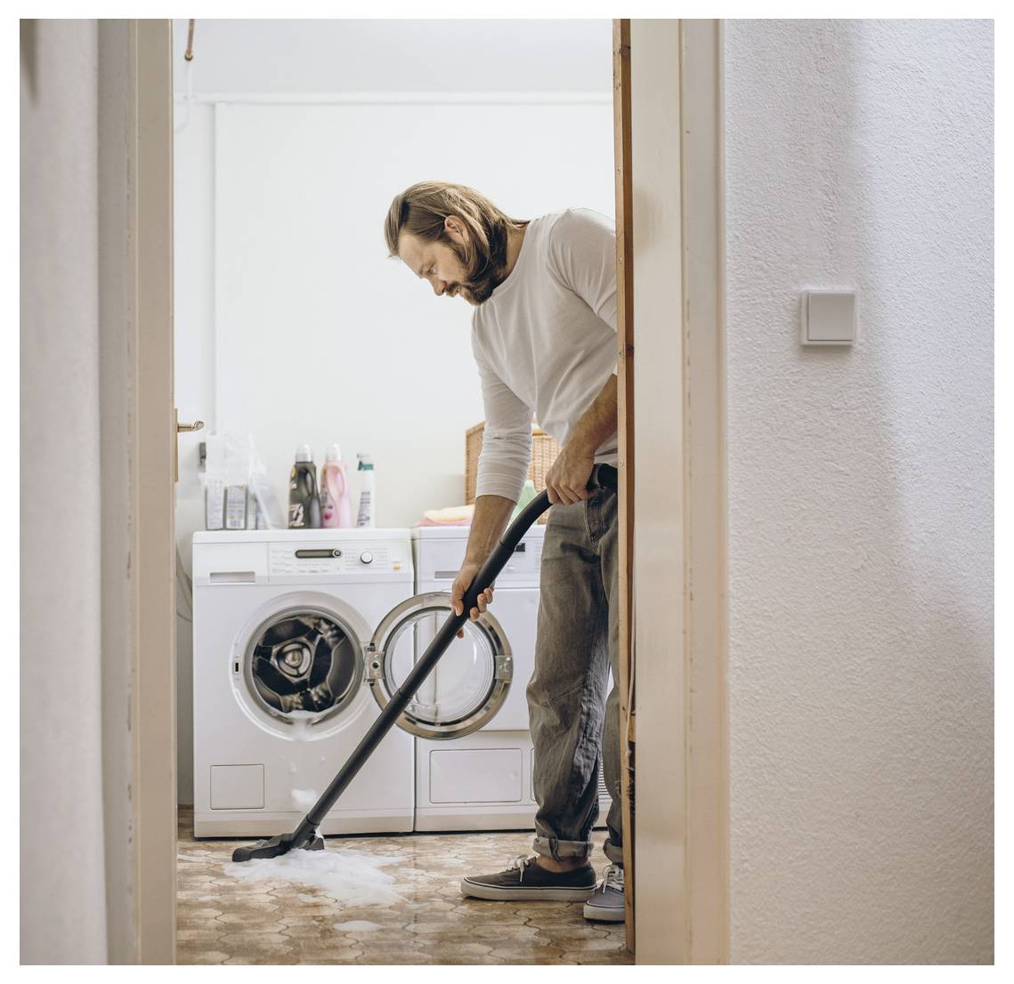 Eine Person saugt den Boden in einem Waschraum, wobei eine Waschmaschine und Reinigungsmittel im Hintergrund zu sehen sind.