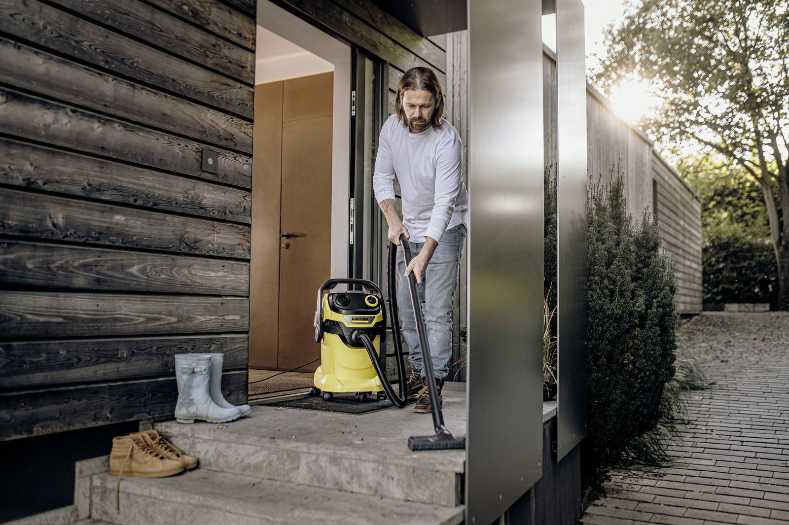 Eine Person saugt die Betonstufen vor einem modernen Holzhaus mit einem tragbaren Staubsauger. In der Nähe stehen Stiefel und die Sonne geht unter.