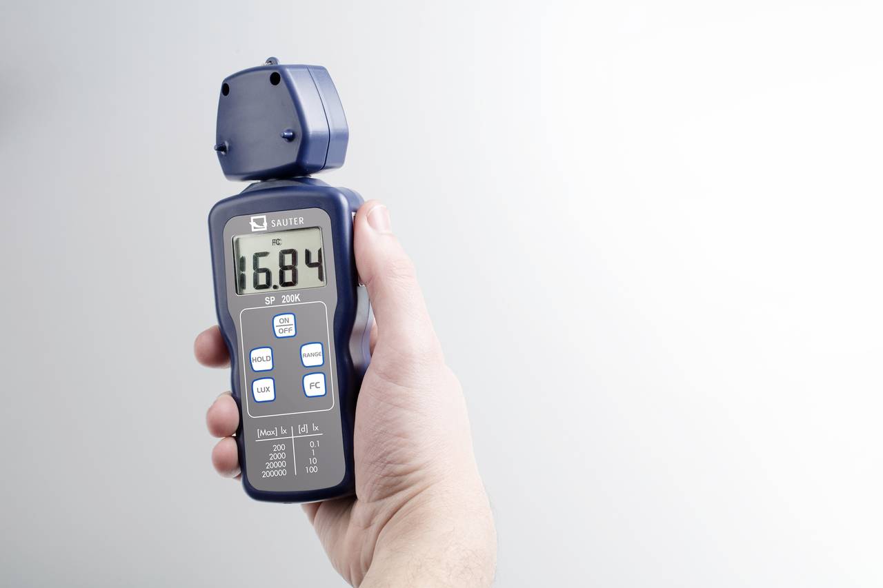 Eine Hand hält ein digitales Anemometer, das eine Anzeige von 16,84 zeigt. Das Gerät misst Windgeschwindigkeit und Temperatur.