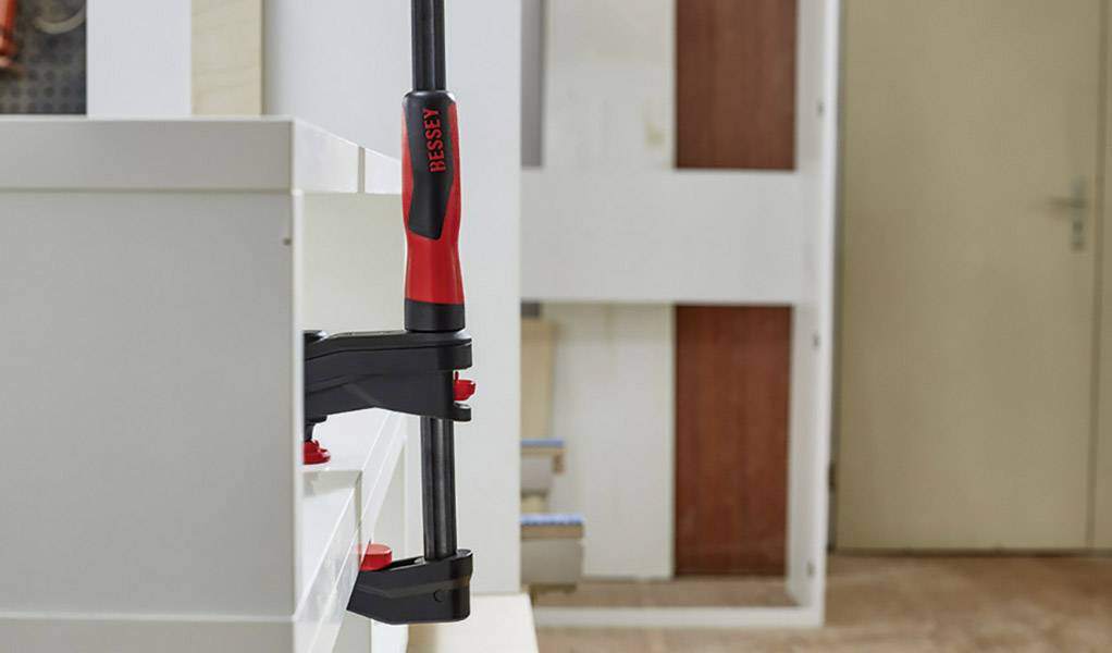 Ein Schraubzwinge von Bessey ist an einem hellen Holzstück befestigt. Im Hintergrund sind ein Türrahmen und ein weiteres Möbelstück zu sehen.