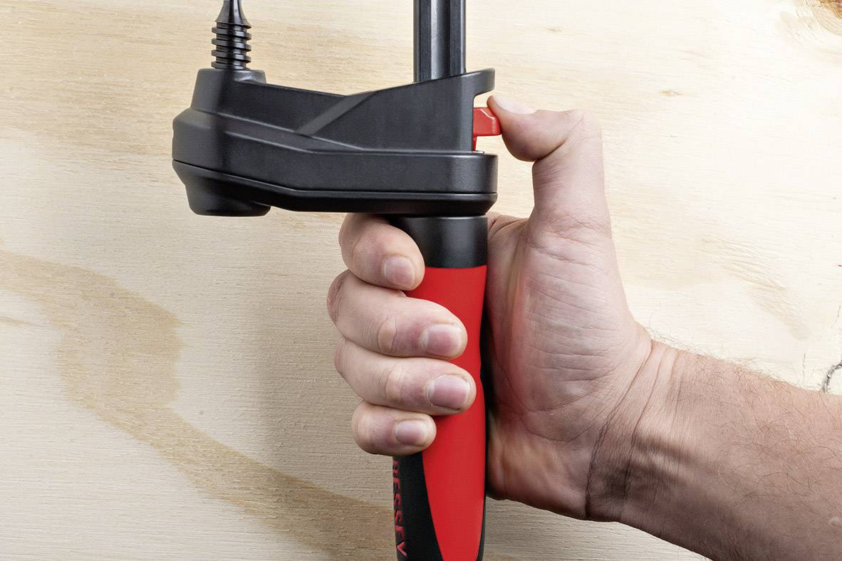 Eine Hand hält einen roten und schwarzen Griff mit einem schwarzen Spannmechanismus an einem Stück Holz.