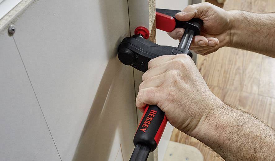 Zwei Hände halten eine rote Zwinge, die verwendet wird, um zwei Holzplatten zusammenzudrücken, auf einem Holzboden.
