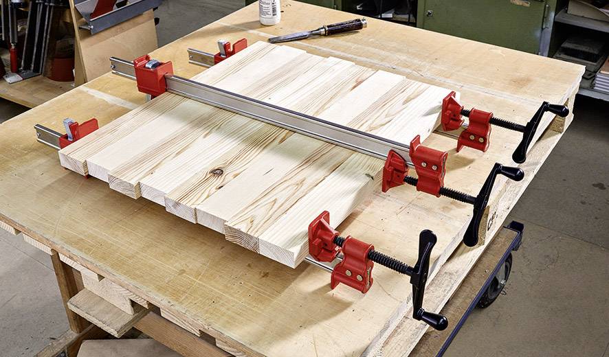 Holzbretter auf Werkbank mit roten Schraubzwingen fixiert, um Verbindung am Stoß zu verleimen. Werkstattumgebung im Hintergrund.