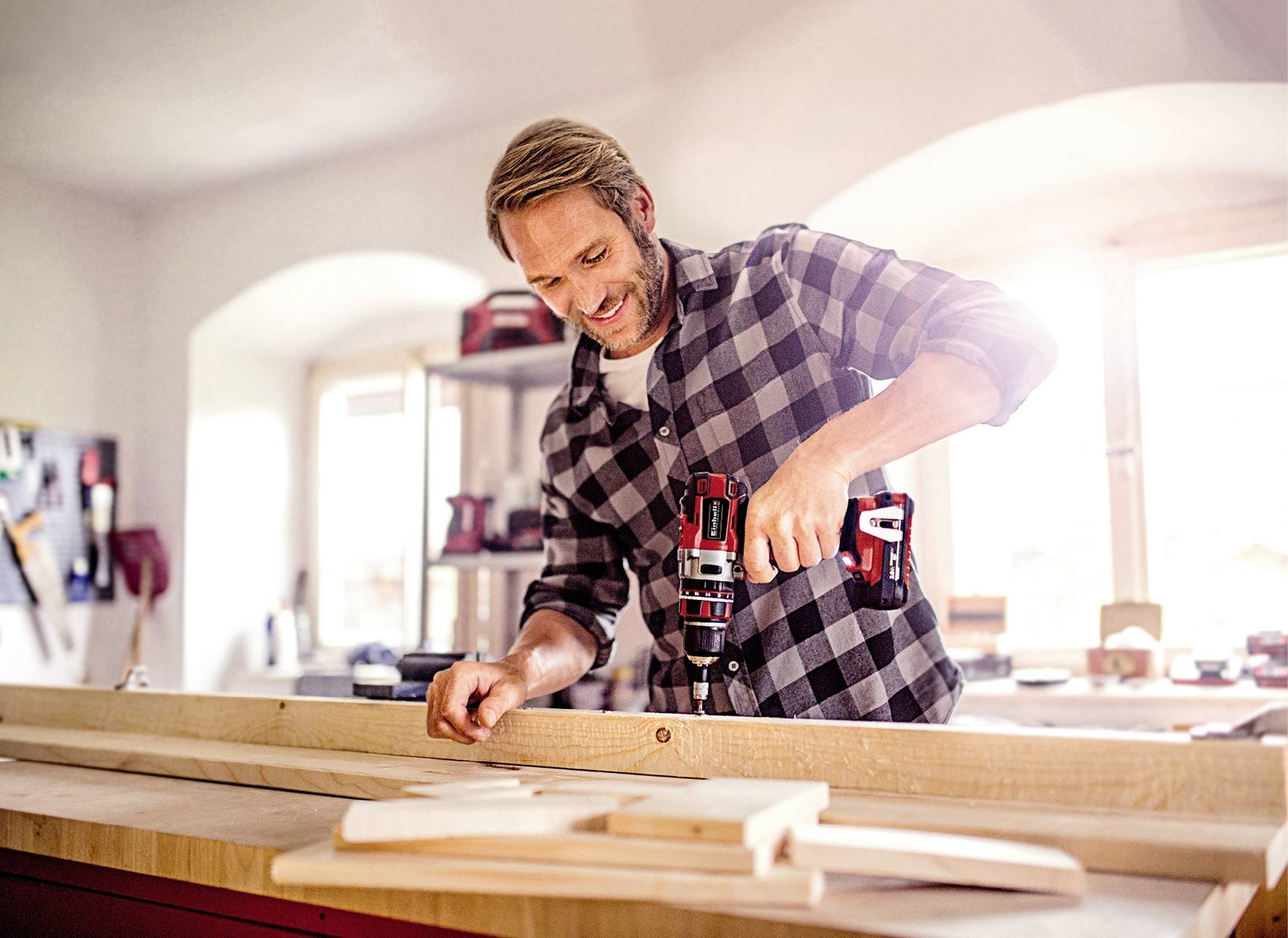 Ein Mann in einem Holzwerkstatt, bohrt mit einer elektrischen Bohrmaschine in ein Stück Holz. Hell beleuchteter Raum im Hintergrund.