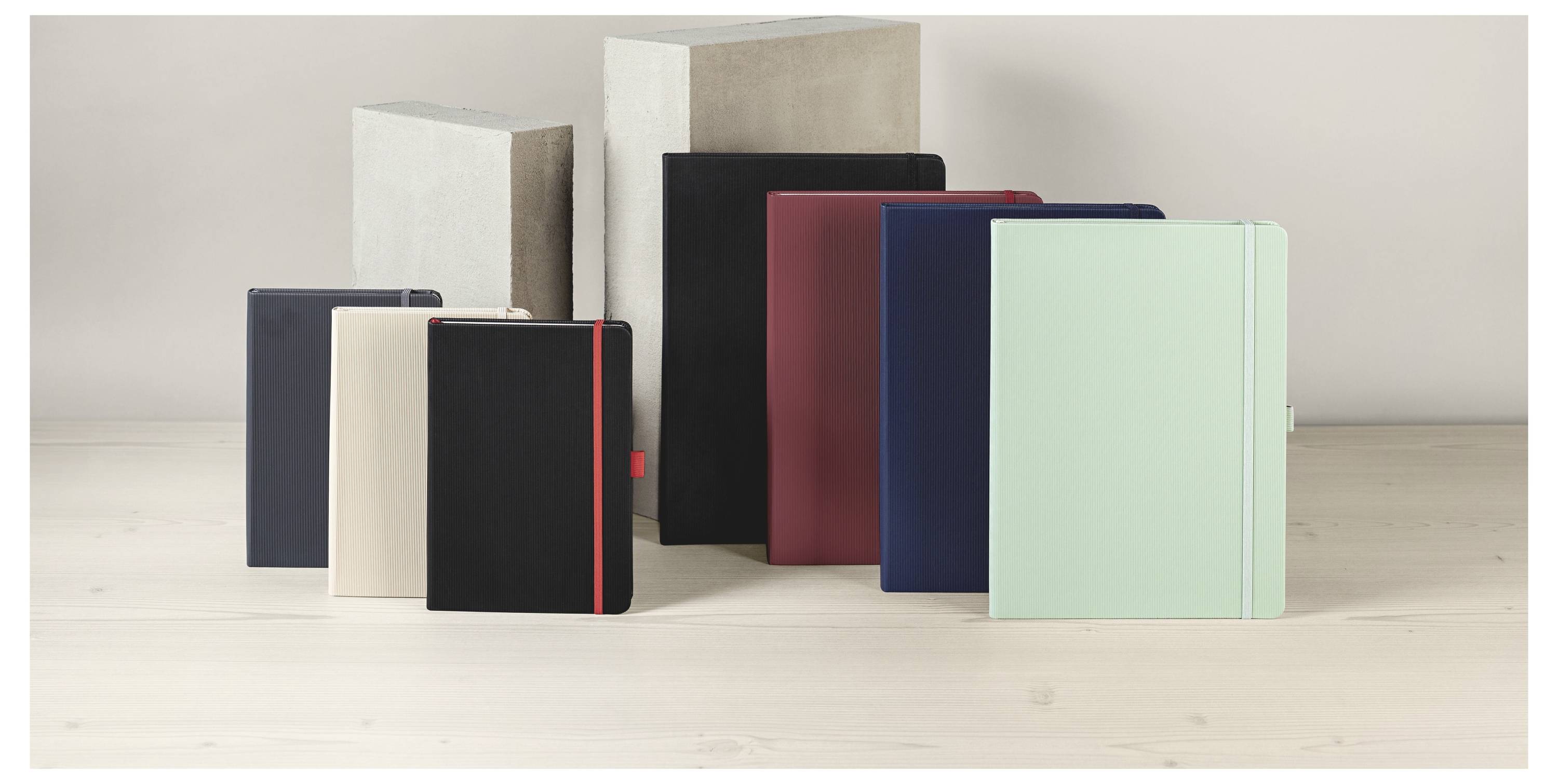 Sortierte gebundene Notizbücher und Journale in Pastelltönen und dunklen Farben, aufrecht auf einer hellen Holzoberfläche vor einem neutralen Hintergrund angeordnet.