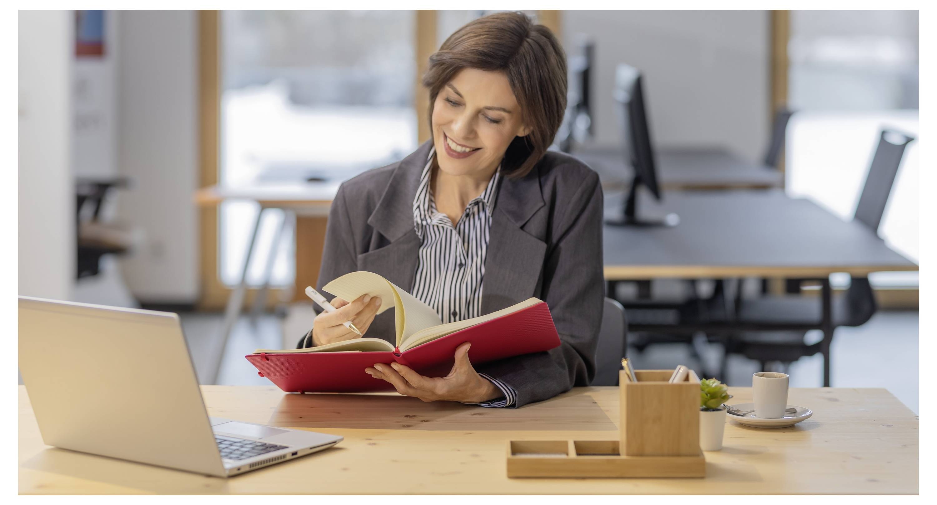 Frau lächelt, während sie in ein rotes Notizbuch an einem Schreibtisch mit einem Laptop in einem Büro schreibt.
