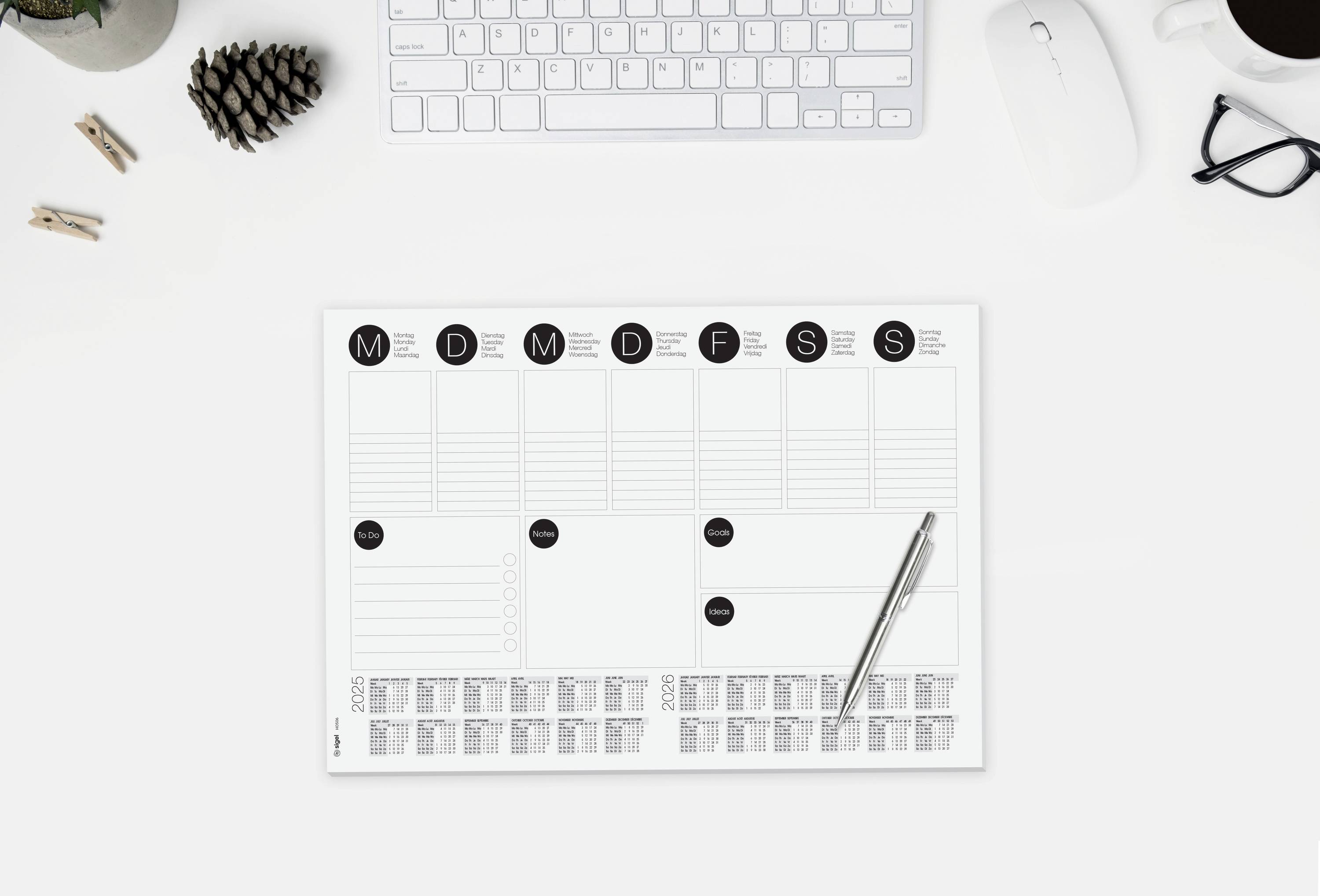 Ein Schreibtisch mit einem Wochenplaner, Stift, Tastatur, Maus, Brille, Tasse Kaffee und einem kleinen Kiefernzapfen.