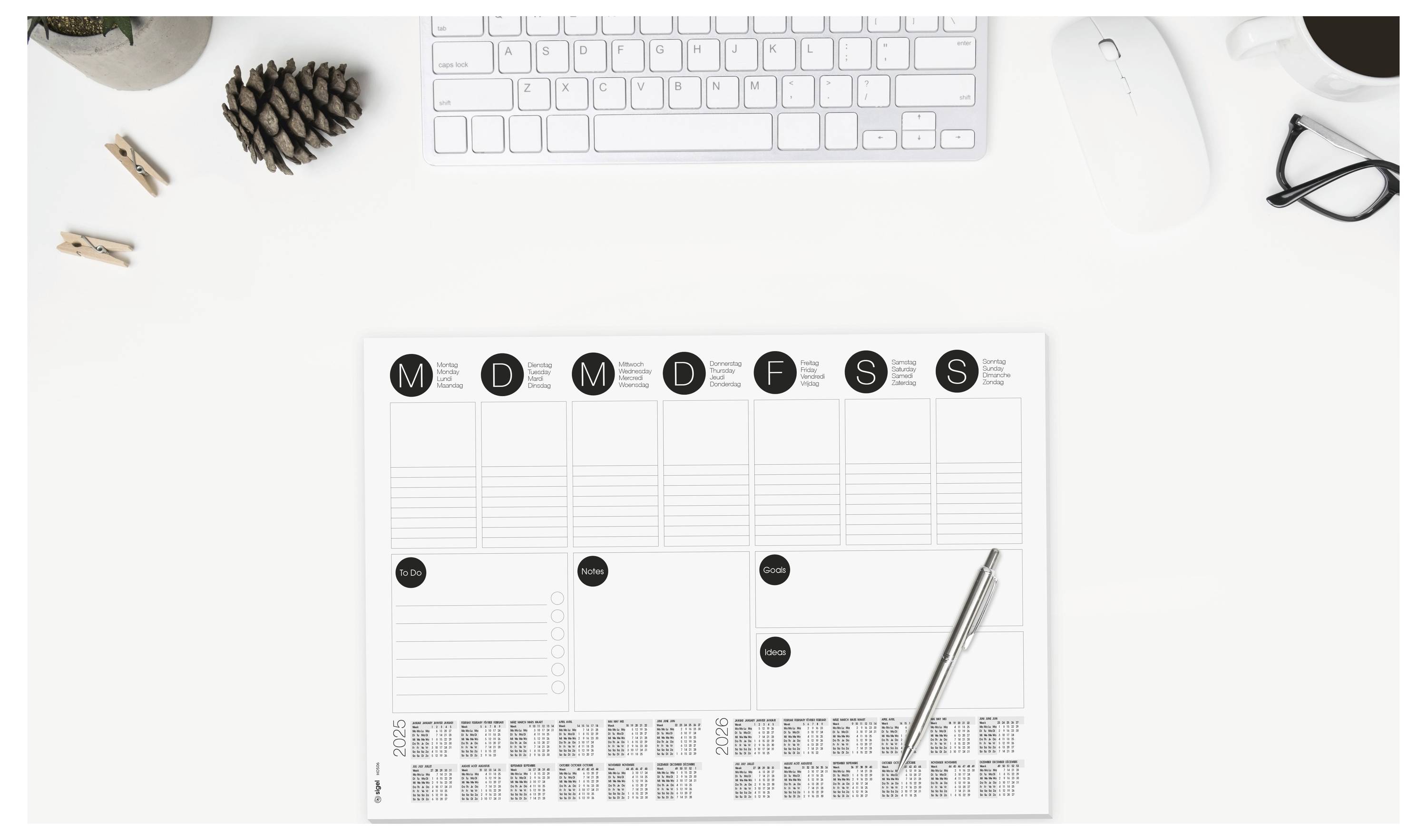 Ansicht von oben auf einen weißen Schreibtisch mit Tastatur, Maus, Brille und Tannenzapfen; ein wöchentlicher Planungsblock mit Spalten, die mit M D M D F S S beschriftet sind, und ein Stift.