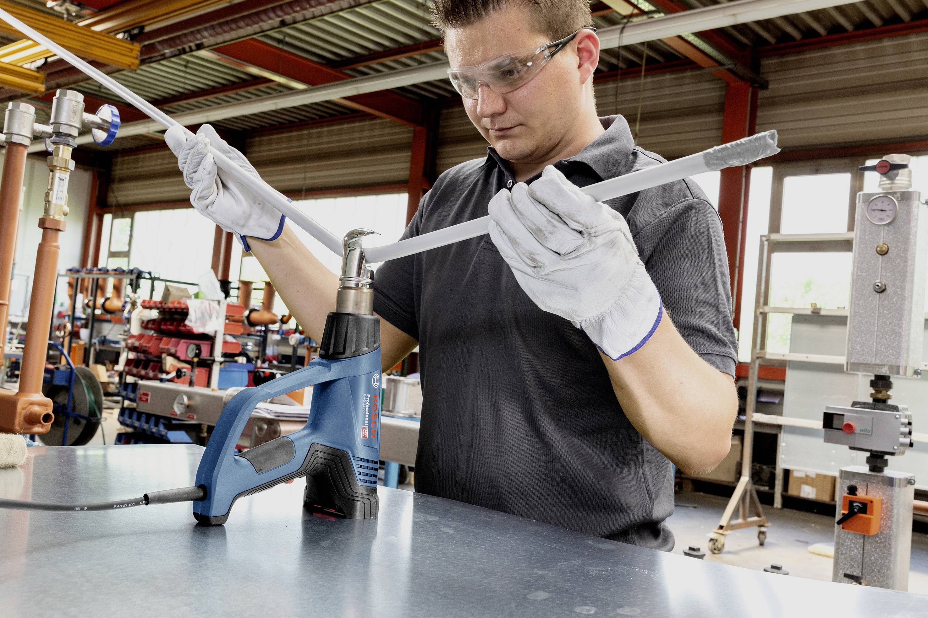 Ein Mann in Arbeitskleidung benutzt Heißluftgebläse und Handschuhe, um ein Rohr in einer Werkstatt zu bearbeiten.