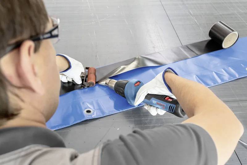 Eine Person verwendet einen Heißluftfön und einen Roller, um eine Naht in Dachbaumaterial zu schweißen, trägt Sicherheitshandschuhe und Schutzbrille auf einer grauen Oberfläche.