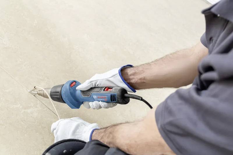 Eine Person verwendet einen blauen Heißluftföhn, um Farbe in einem Raum zu entfernen. Sie trägt Handschuhe und ein dunkles Shirt und konzentriert sich auf die Aufgabe.