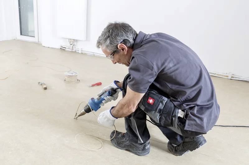 Eine Person kniet auf dem Boden und verwendet eine Bohrmaschine, um an einer Oberfläche zu arbeiten. Werkzeuge und Kabel sind verstreut, was auf eine Reparaturaufgabe hindeutet.