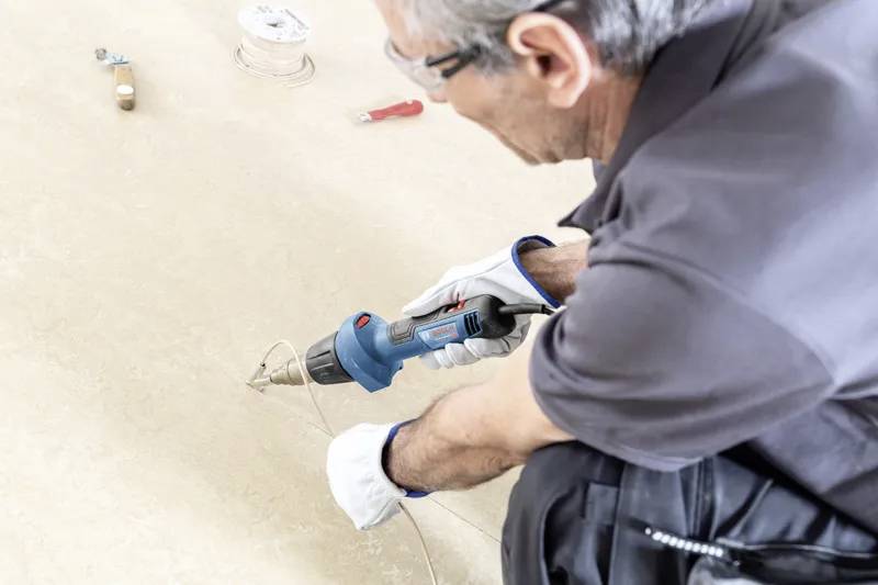 Eine Person mit Handschuhen und Schutzbrille verwendet einen blauen Heißluftfön auf einer Oberfläche, höchstwahrscheinlich für den Einbau oder die Reparatur von Bodenbelägen. Werkzeuge liegen in der Nähe.