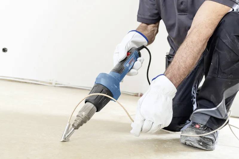 Eine Person verwendet einen Heißluftfön an einem Kabel, das auf dem Boden liegt, und trägt dabei Schutzhandschuhe und Arbeitskleidung, konzentriert auf eine Bauaufgabe.
