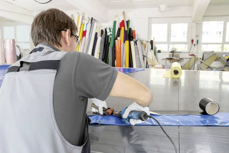 Ein Arbeiter verwendet ein Werkzeug, um zwei Blätter aus blauem Material in einer Fertigungswerkstatt zu versäubern, umgeben von Rollen bunter Materialien.