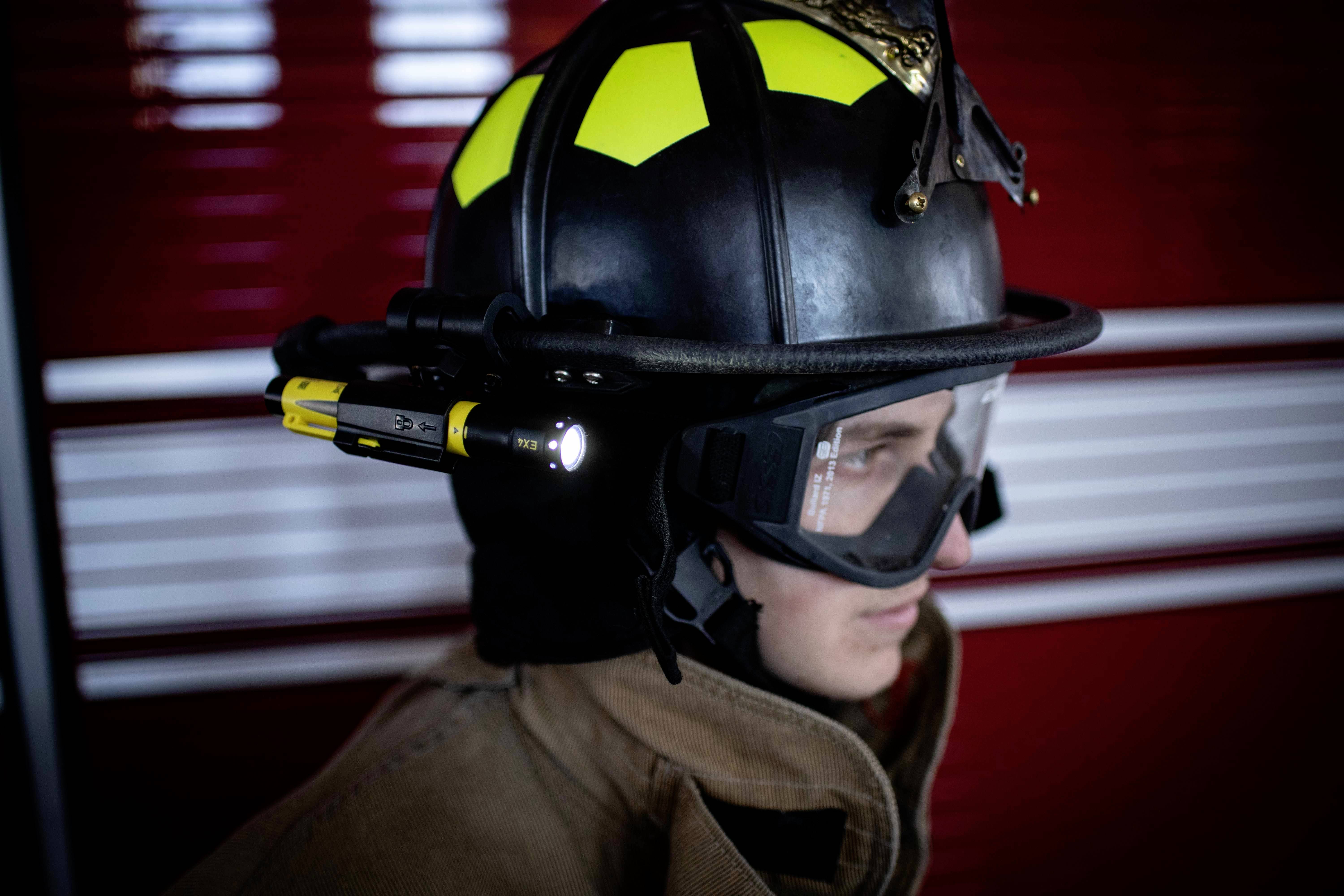 Ein Feuerwehrmann im Profil trägt einen Helm mit integrierter Lampe, Schutzbrille und Schutzanzug vor einem roten Hintergrund.