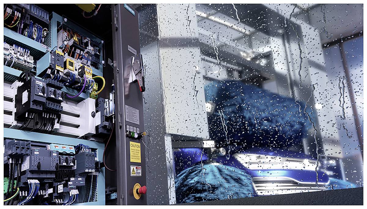 Industrieanlage hinter regennassem Fenster, zeigt Schaltkasten links mit Kabeln, Schaltern und Bauteilen; Produktionsmaschine im Hintergrund.