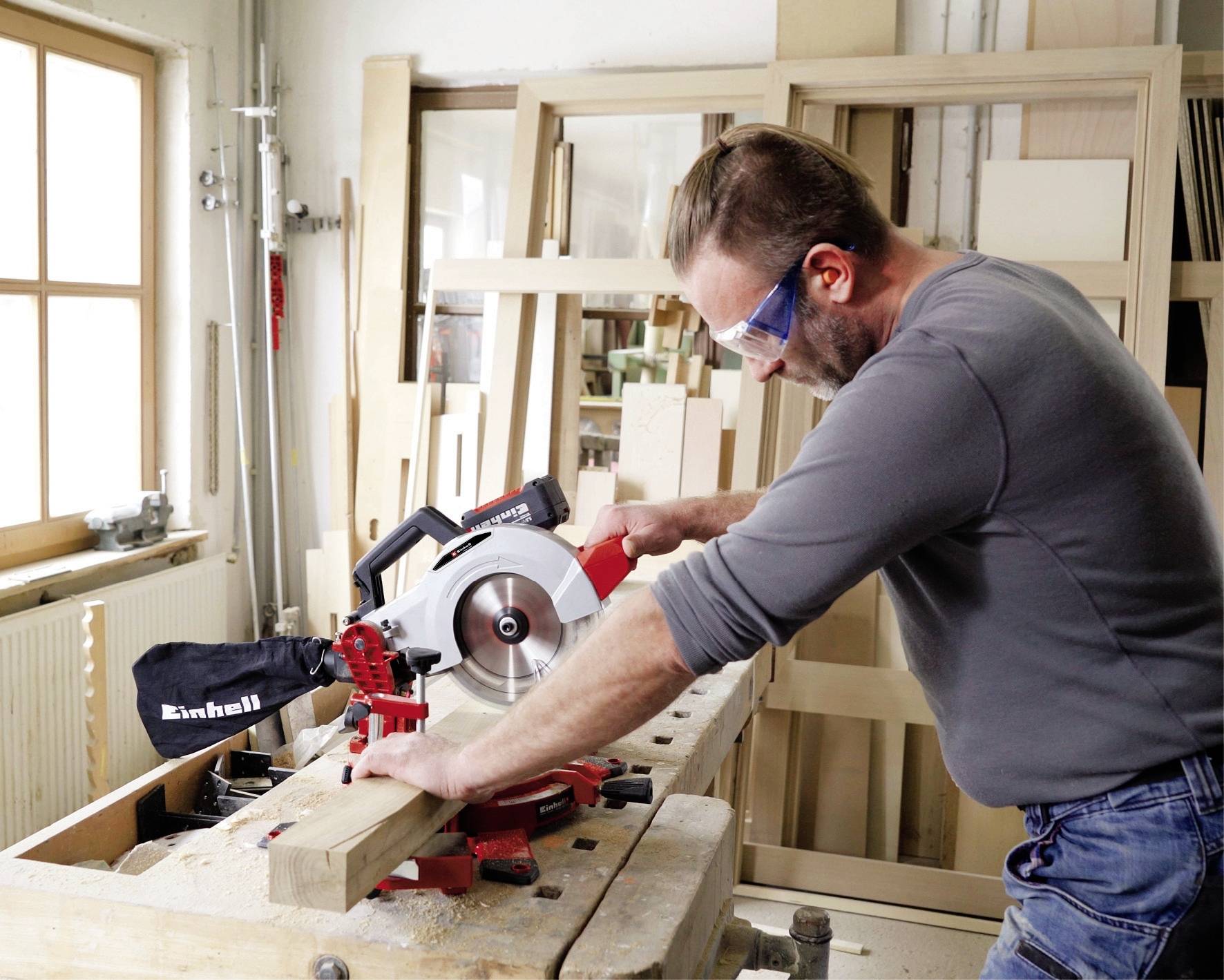 Ein Mann in grauem Oberteil arbeitet mit einer Tischsäge in einer Werkstatt. Holzstücke und Rahmen sind im Hintergrund zu sehen.