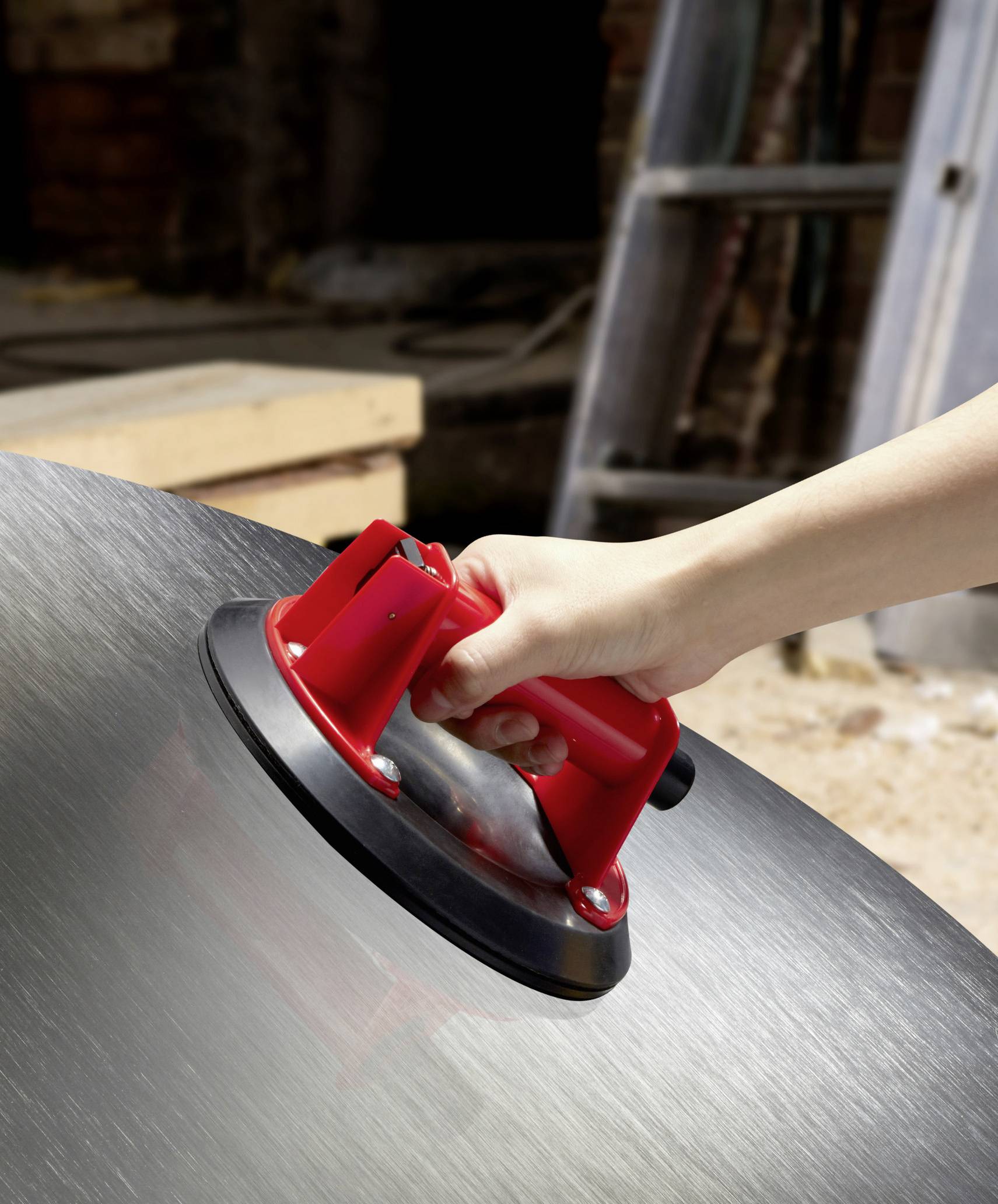 Hand benutzt einen roten Saugnapf, um eine Metallplatte anzuheben. Baustelle im Hintergrund mit unscharfen Werkzeugen und Materialien.