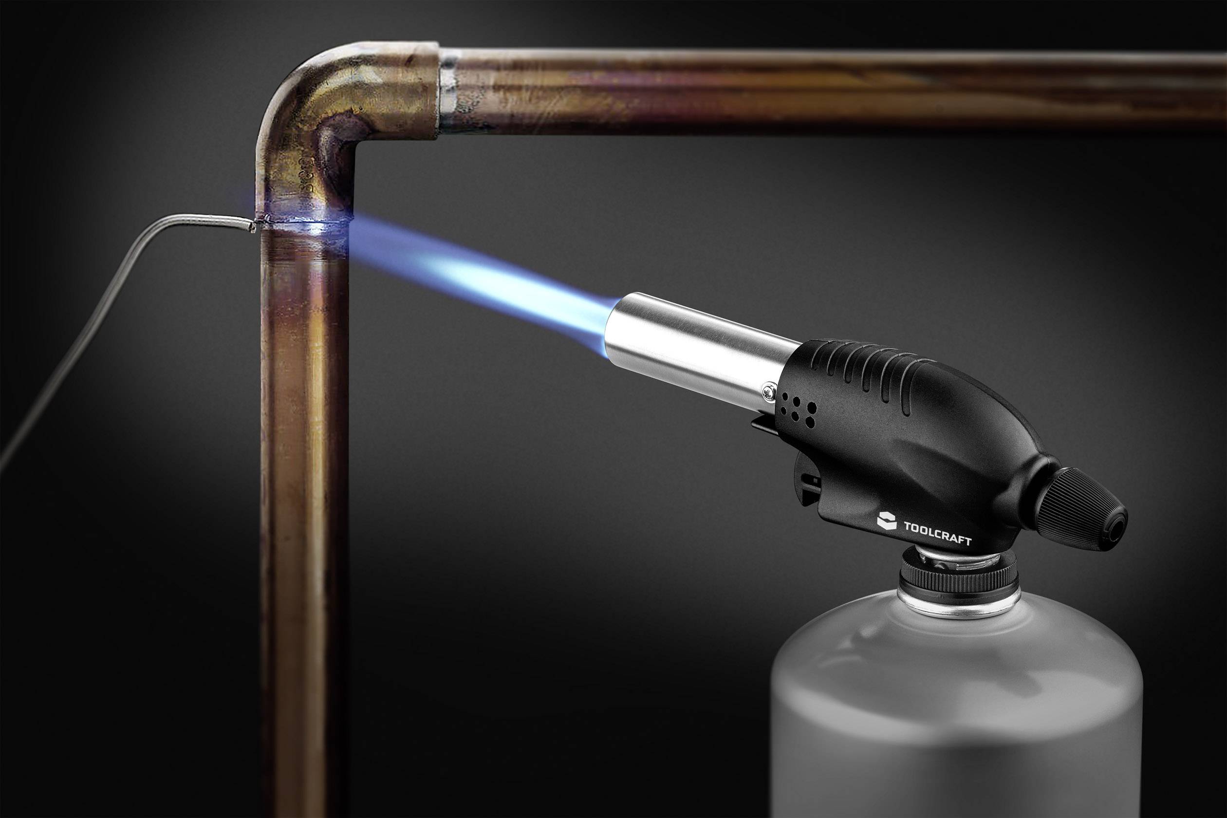 Ein Gasbrenner erhitzt ein Kupferrohr an der Biegung für eine Lötverbindung, blauer Flammenstrahl deutlich sichtbar.