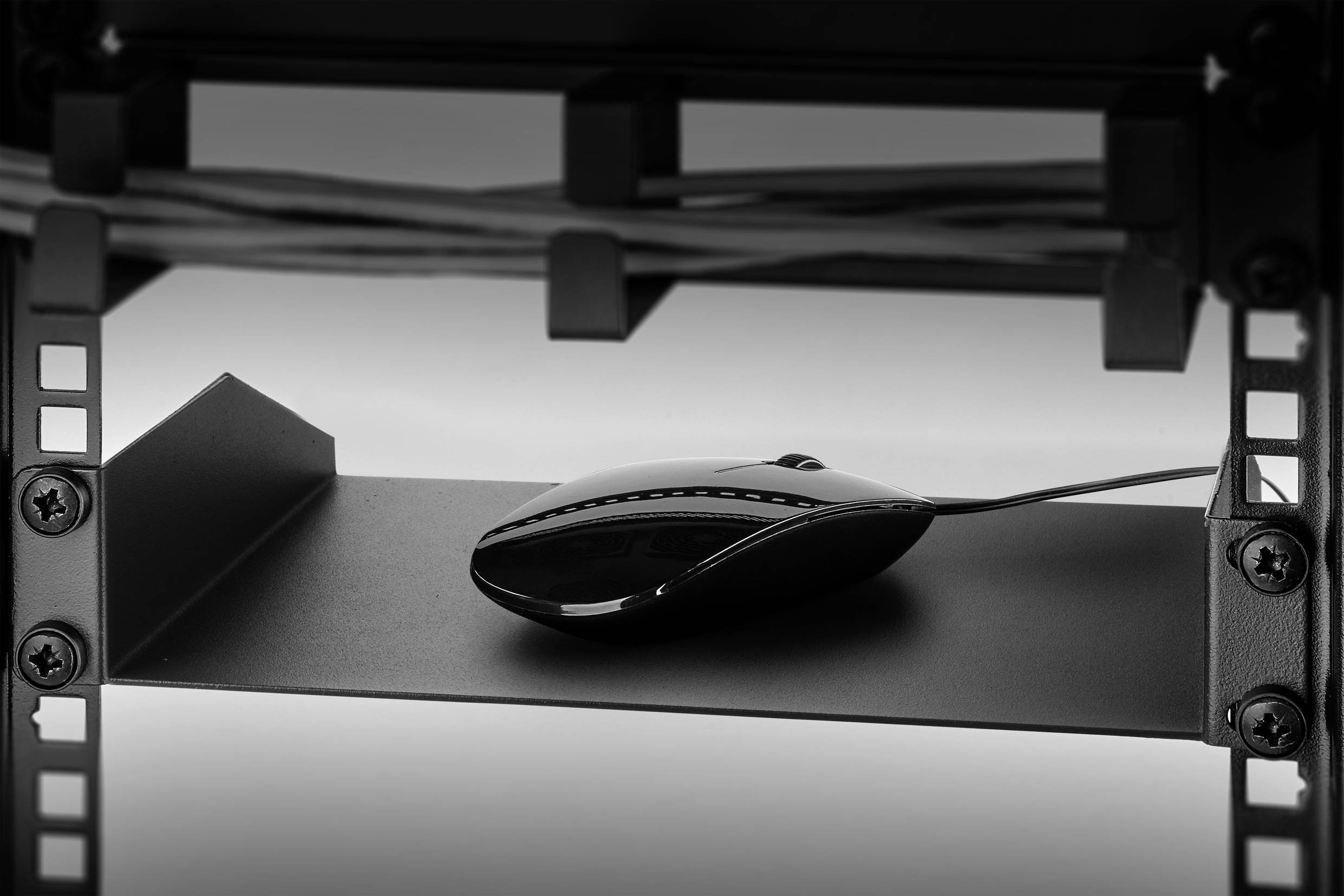Eine schwarze Computermaus liegt auf einem metallischen Regalboden unter einem Tisch mit Kabeln im Hintergrund.