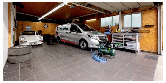 Garageninneres mit einem weißen Transporter und einem Oldtimer, die geparkt sind. Ein Kompressor mit einem Schlauch liegt in der Nähe des Transporters. Werkzeuge sind auf Regalen organisiert.