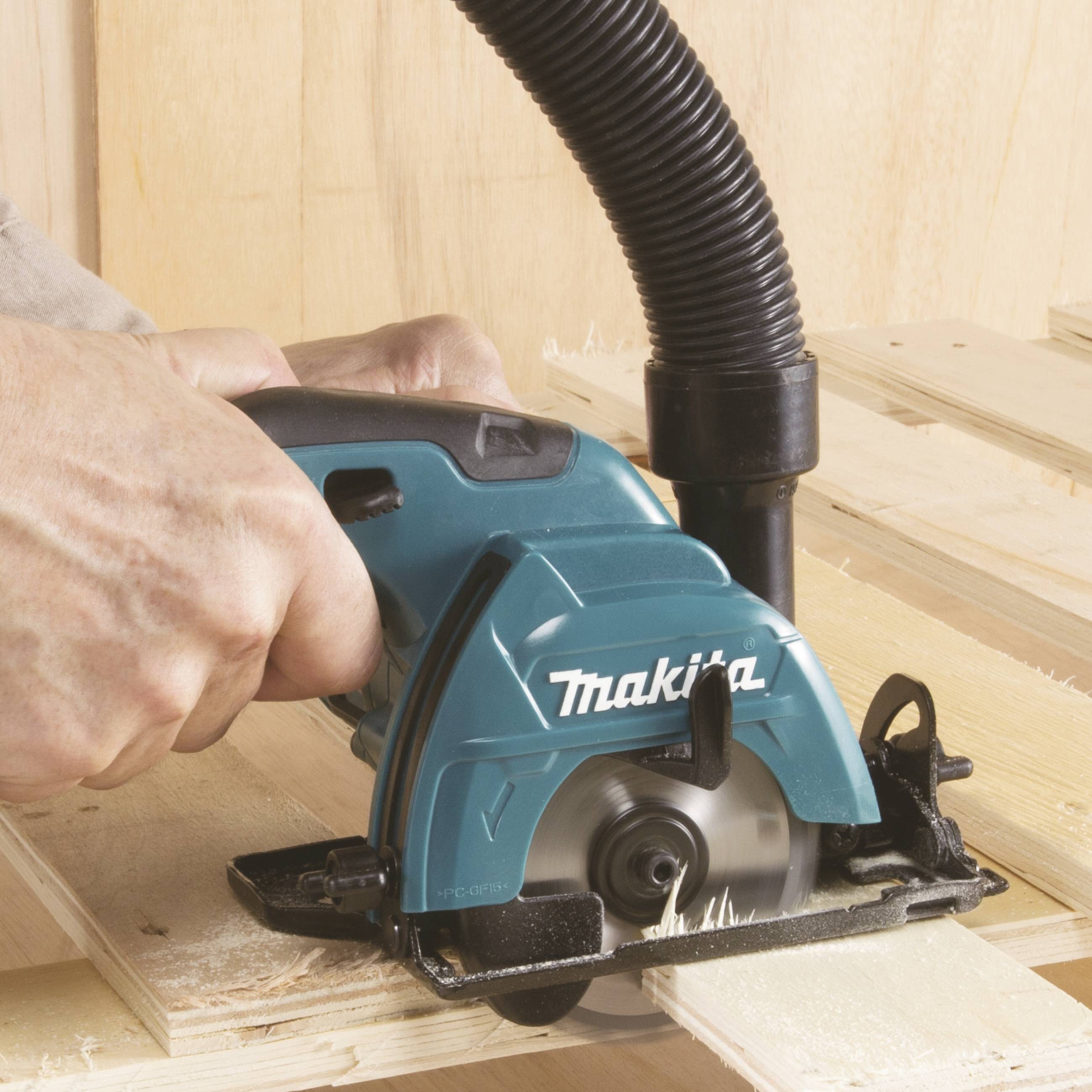 Eine Hand hält eine Elektrosäge, die Holz schneidet. Staub wird von einem angeschlossenen Schlauch abgesaugt.