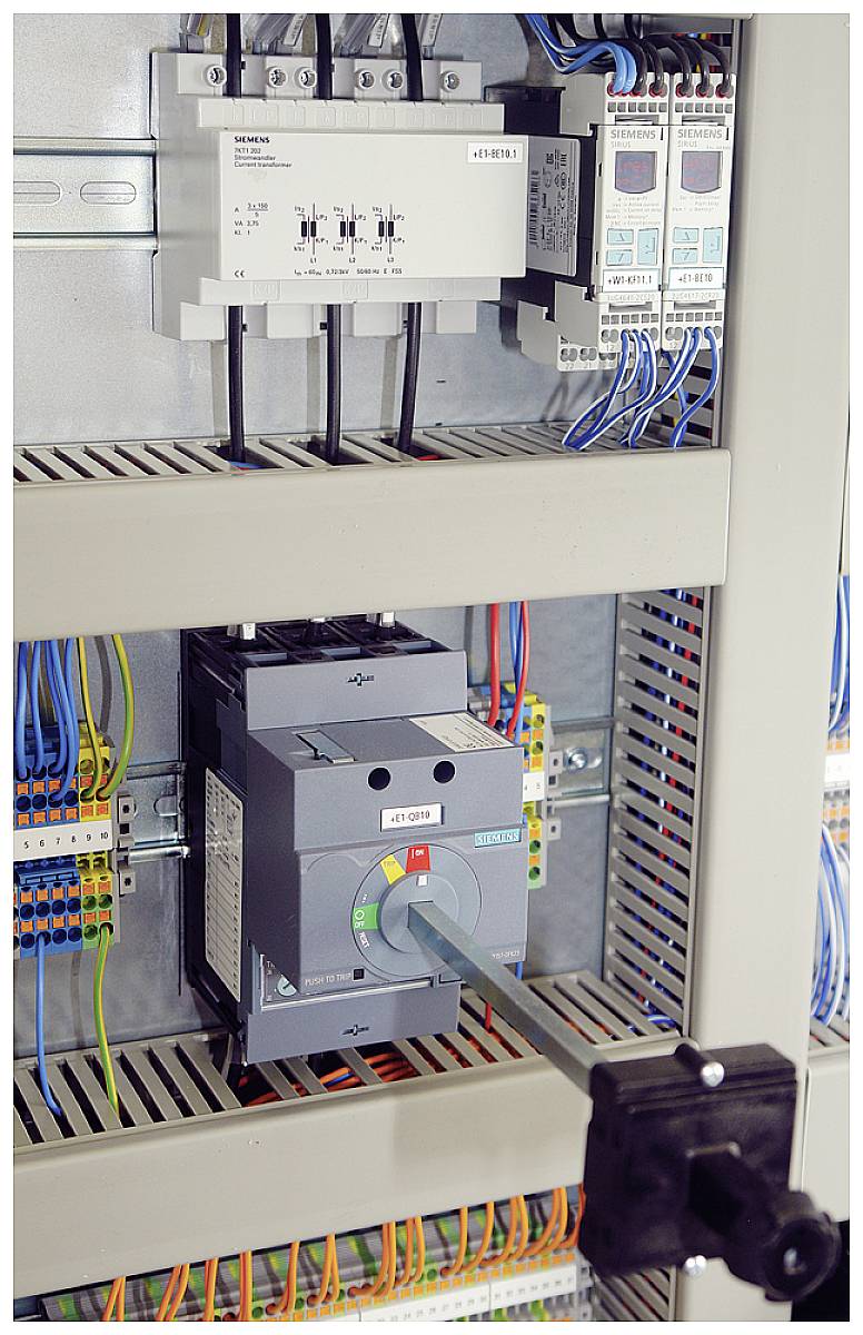 Ein geöffneter Schaltschrank zeigt verkabelte elektrische Komponenten und Sicherungen, mit einem Hauptschalter in der Mitte.