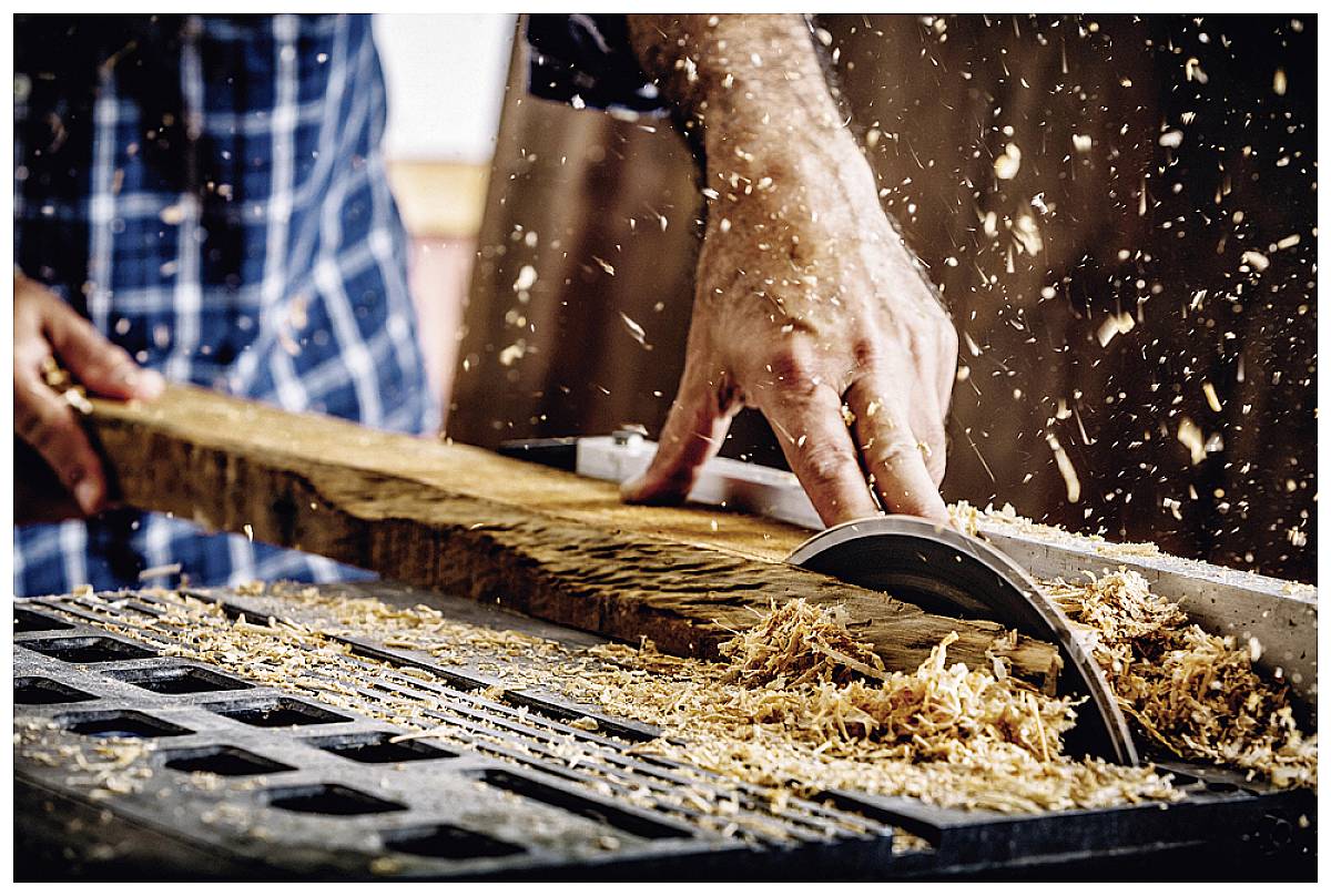 Ein Handwerker schneidet ein Holzbrett mit einer Kreissäge. Holzspäne fliegen in der Luft. Die Person trägt ein kariertes Hemd.