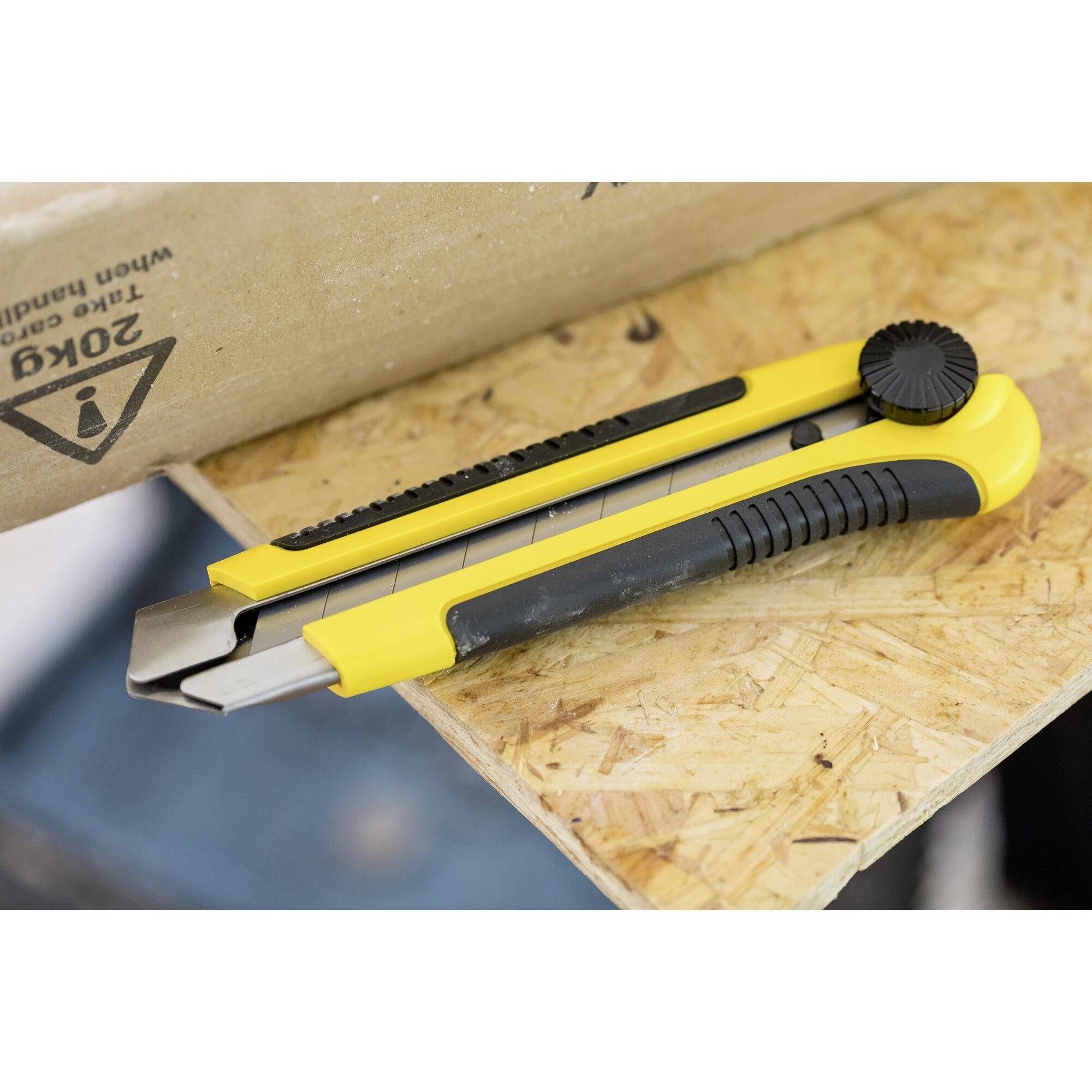 Ein gelber Cutter liegt auf einer Holzplatte neben einem Karton. Der Cutter hat einen schwarzen Griff und wird für Schneidearbeiten verwendet.