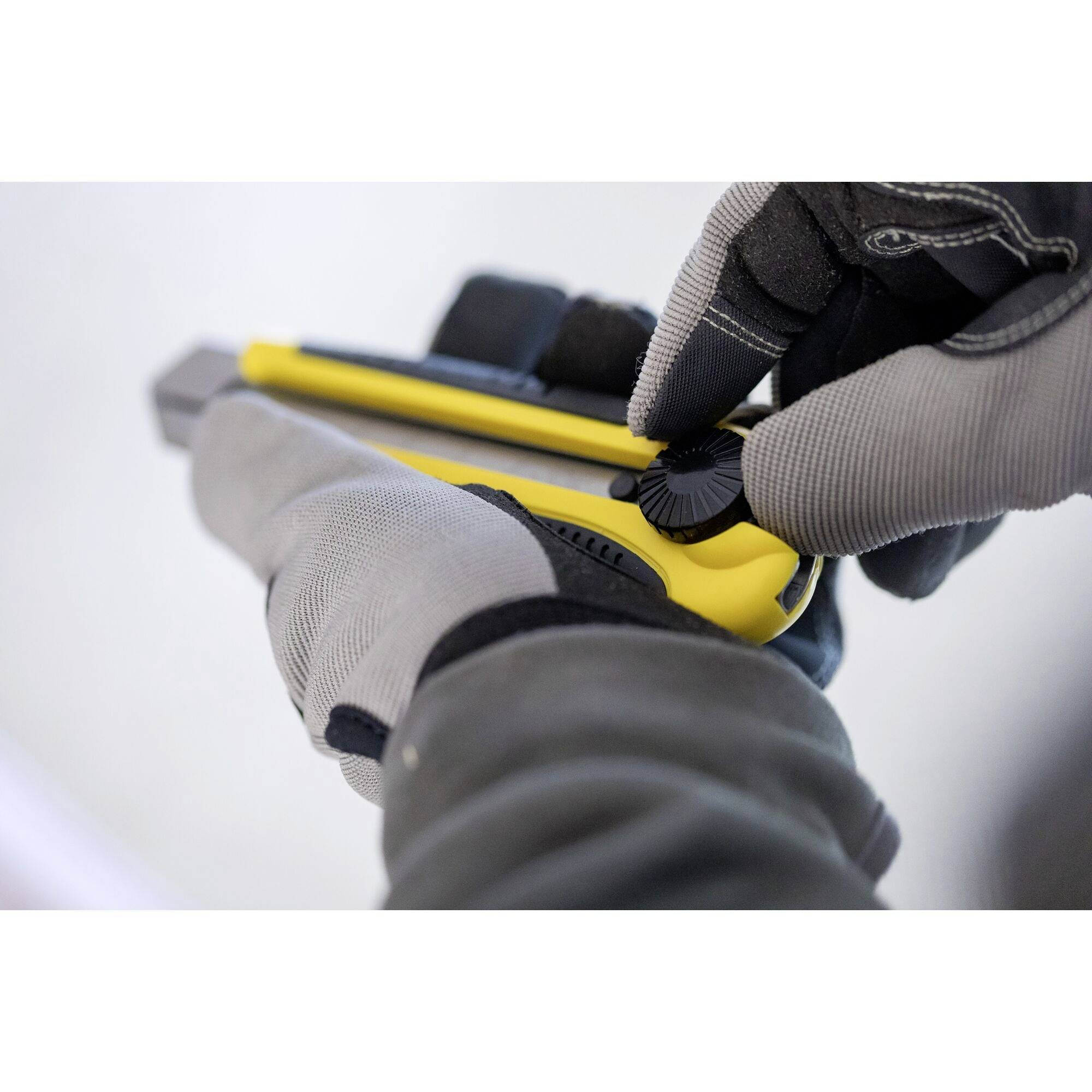 Eine Person mit grauen Handschuhen hält einen gelben Cutter-Messer. Die Hand justiert die Klinge mit einem schwarzen Rad.