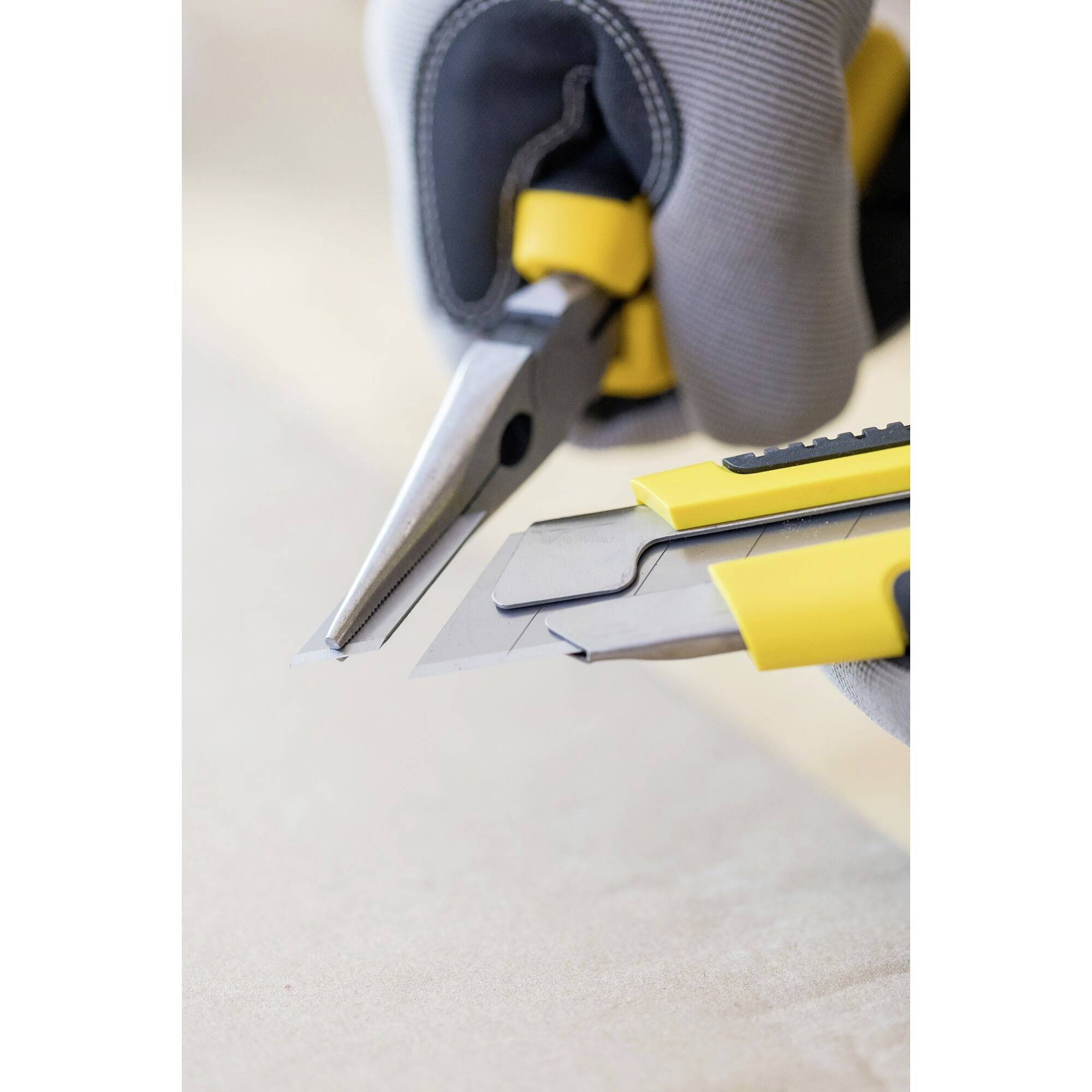 Jemand hält eine Zange und ein Teppichmesser, beide mit gelben Griffen. Die Zange schneidet ein Metallteil, symbolisch für Handarbeit.