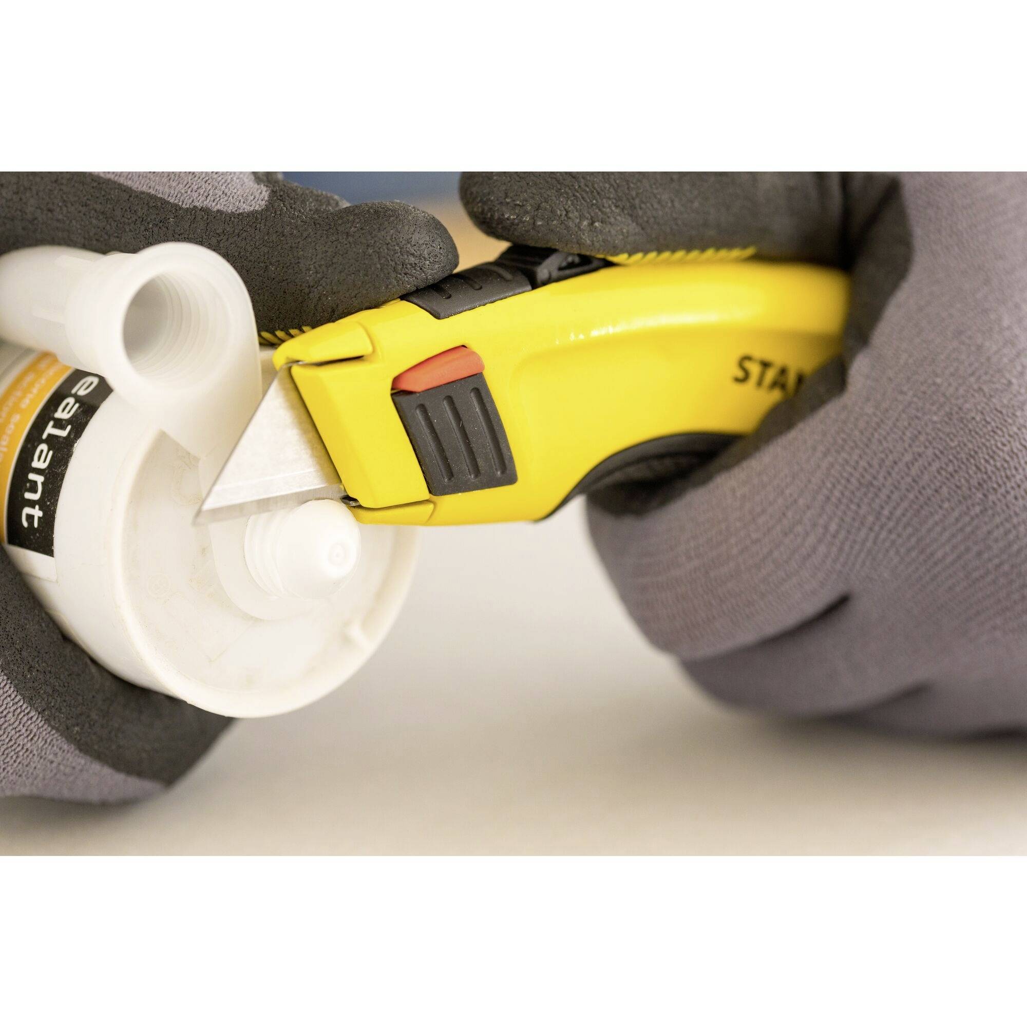 Eine Person, die graue Handschuhe trägt, schneidet die Spitze eines Klebstoffbehälters mit einem gelben Cuttermesser ab.