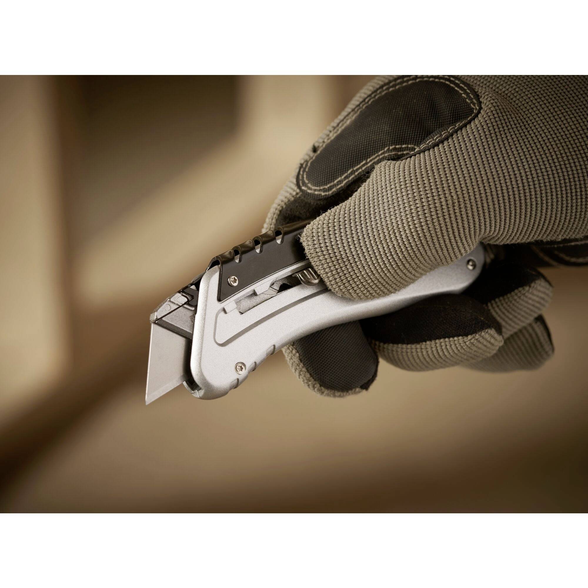 Eine Hand hält ein silberfarbenes Cuttermesser mit schwarzem Griff, bereit zum Schneiden. Die Person trägt einen braunen Arbeitshandschuh.