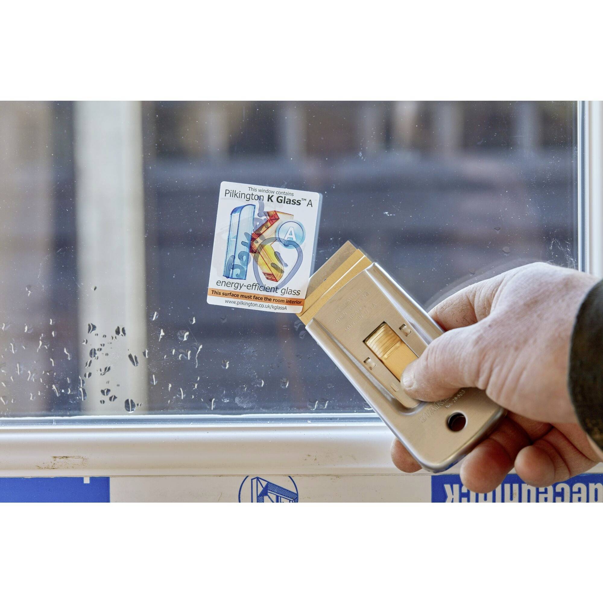 Eine Person entfernt ein Etikett von einem Fenster. Auf dem Etikett steht 'Pilkington K Glass™ A'. Das Fenster zeigt 'energy-efficient glass'.