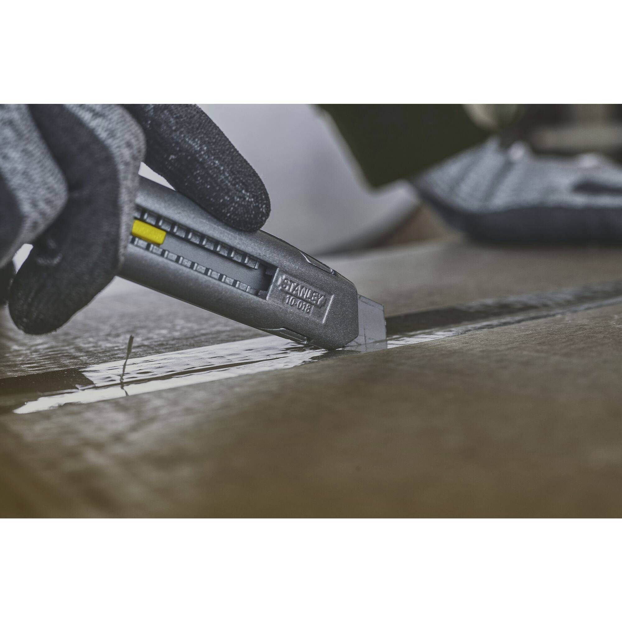 Eine behandschuhte Hand schneidet mit einem Teppichmesser einen Pappkarton auf. Das Messer hat eine Skala an der Seite.