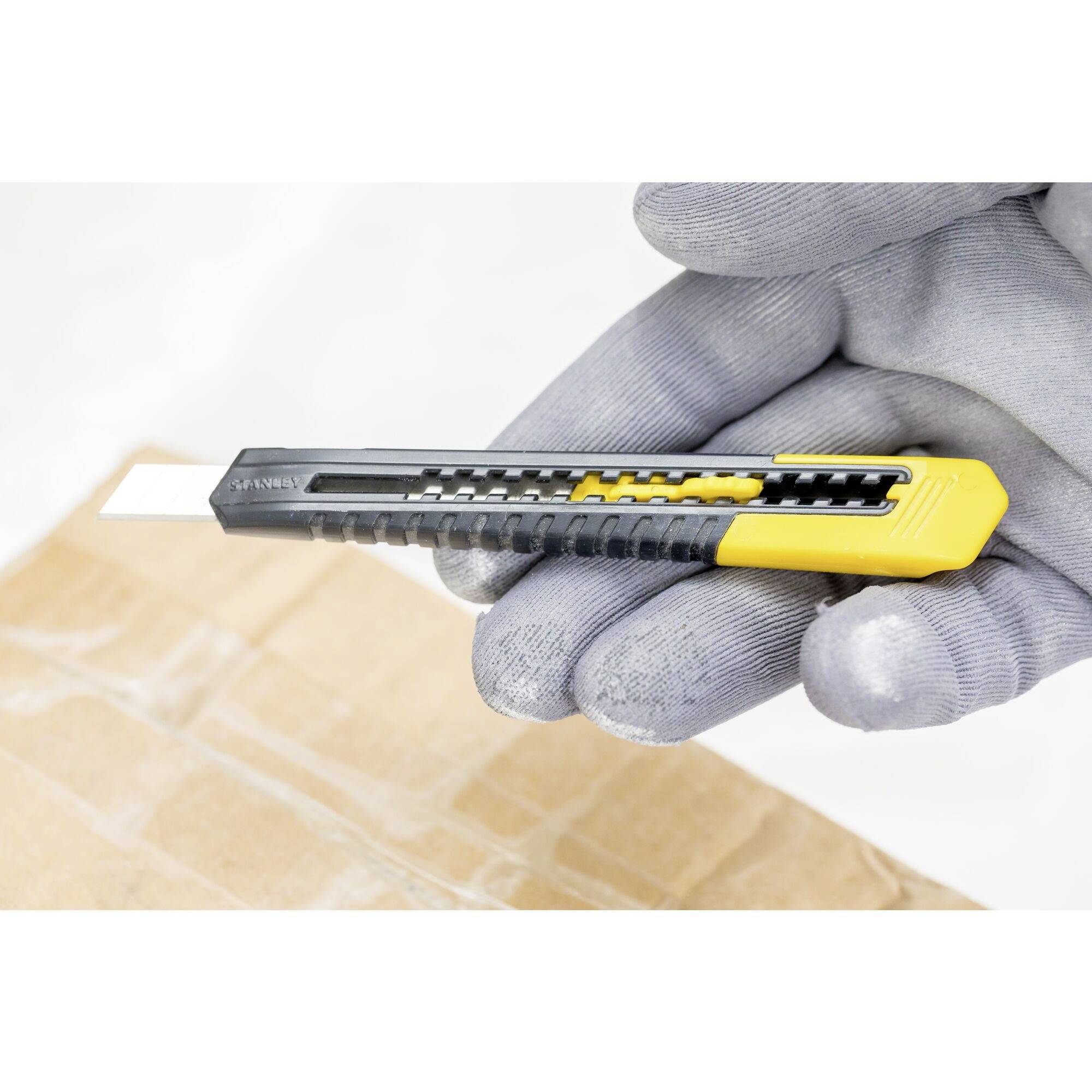 Eine behandschuhte Hand hält ein gelbes Cuttermesser, über einem braunen Karton schwebend, bereit zum Schneiden.