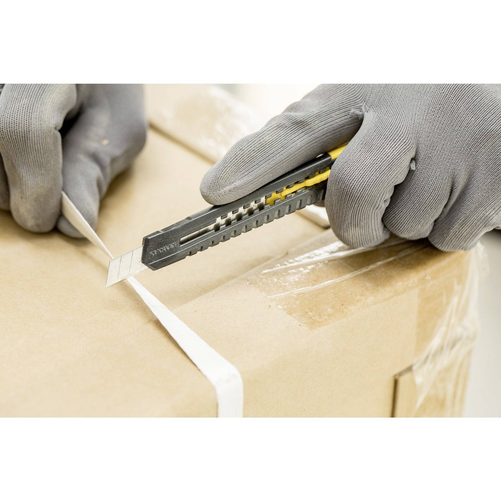 Eine Person öffnet mit einem Cutter-Messer einen Karton. Sie trägt graue Arbeitshandschuhe.