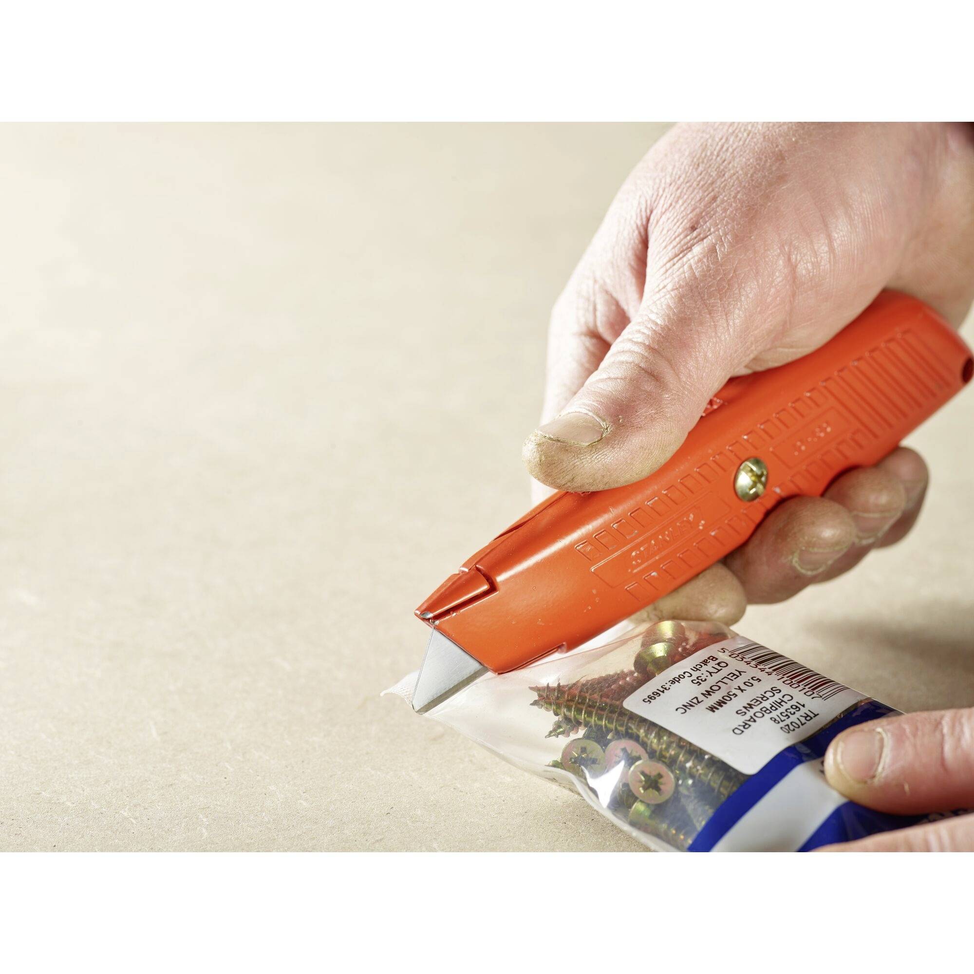 Eine Hand benutzt einen orangefarbenen Kartonöffner, um eine Plastiktüte mit Schrauben zu öffnen. Im Hintergrund ist ein Tisch zu sehen.