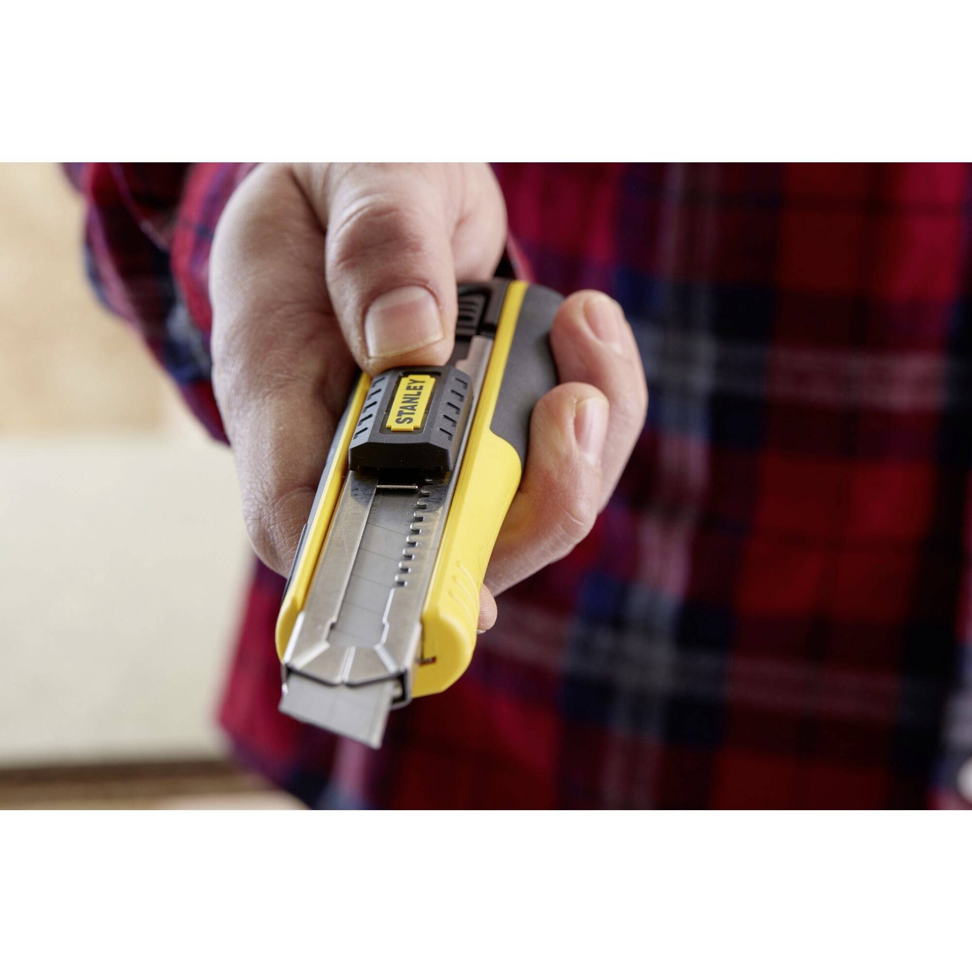 Eine Hand hält ein gelbes Teppichmesser, das bereit zum Schneiden ist. Die Person trägt ein kariertes Hemd.
