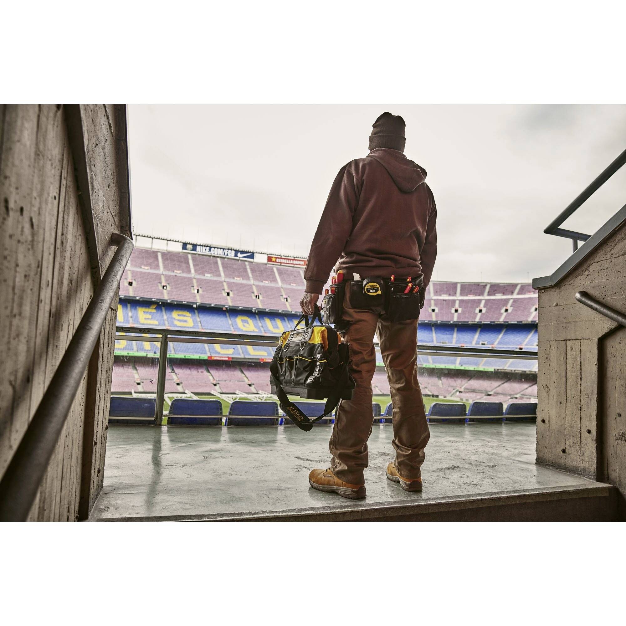 Eine Person in Arbeitskleidung steht mit einer Tasche im Fußballstadion und blickt auf das Spielfeld.