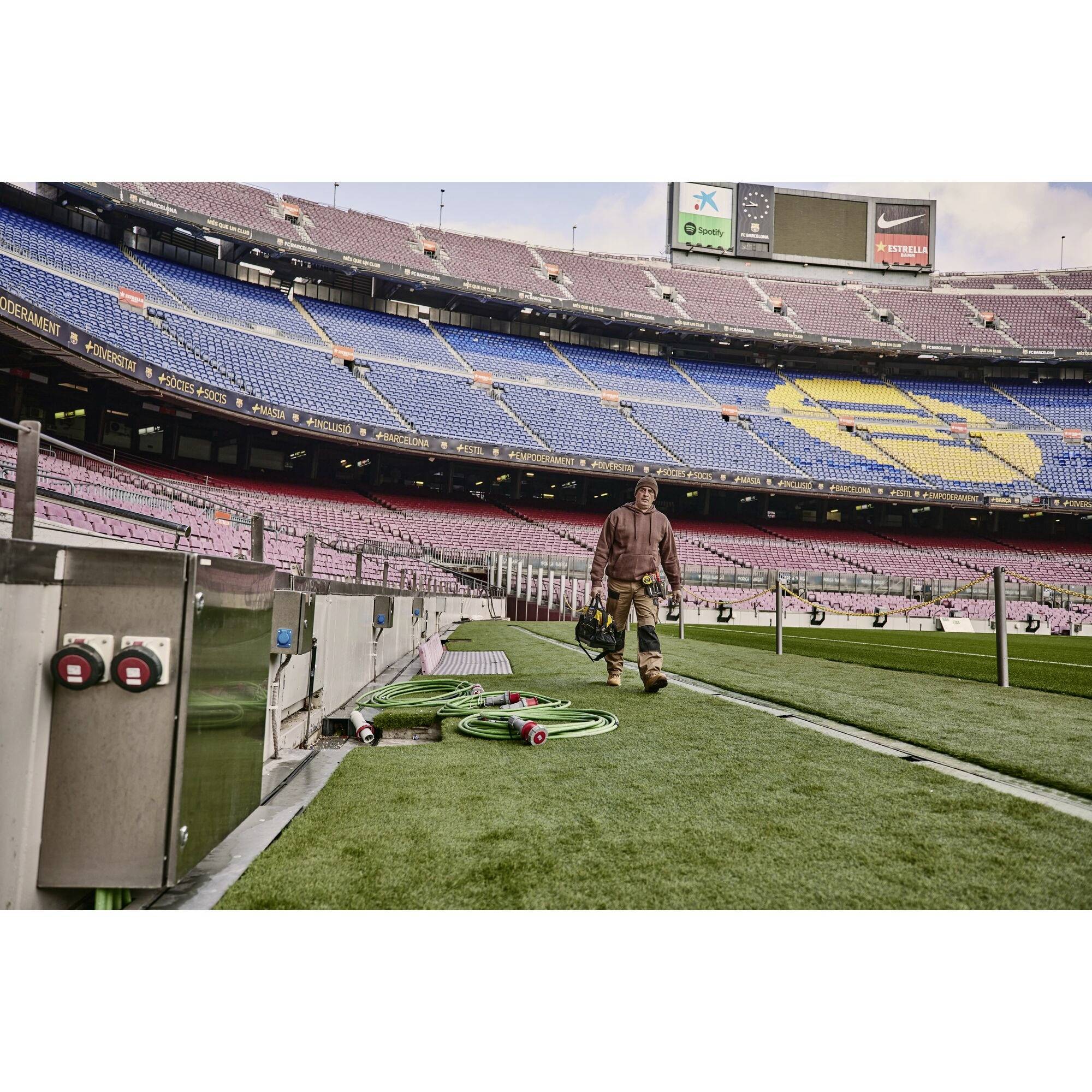 Ein Stadion mit leeren Sitzreihen in Blau und Rot. Ein Arbeiter trägt Ausrüstung über einen grünen Rasen, mehrere Schläuche liegen auf dem Boden.